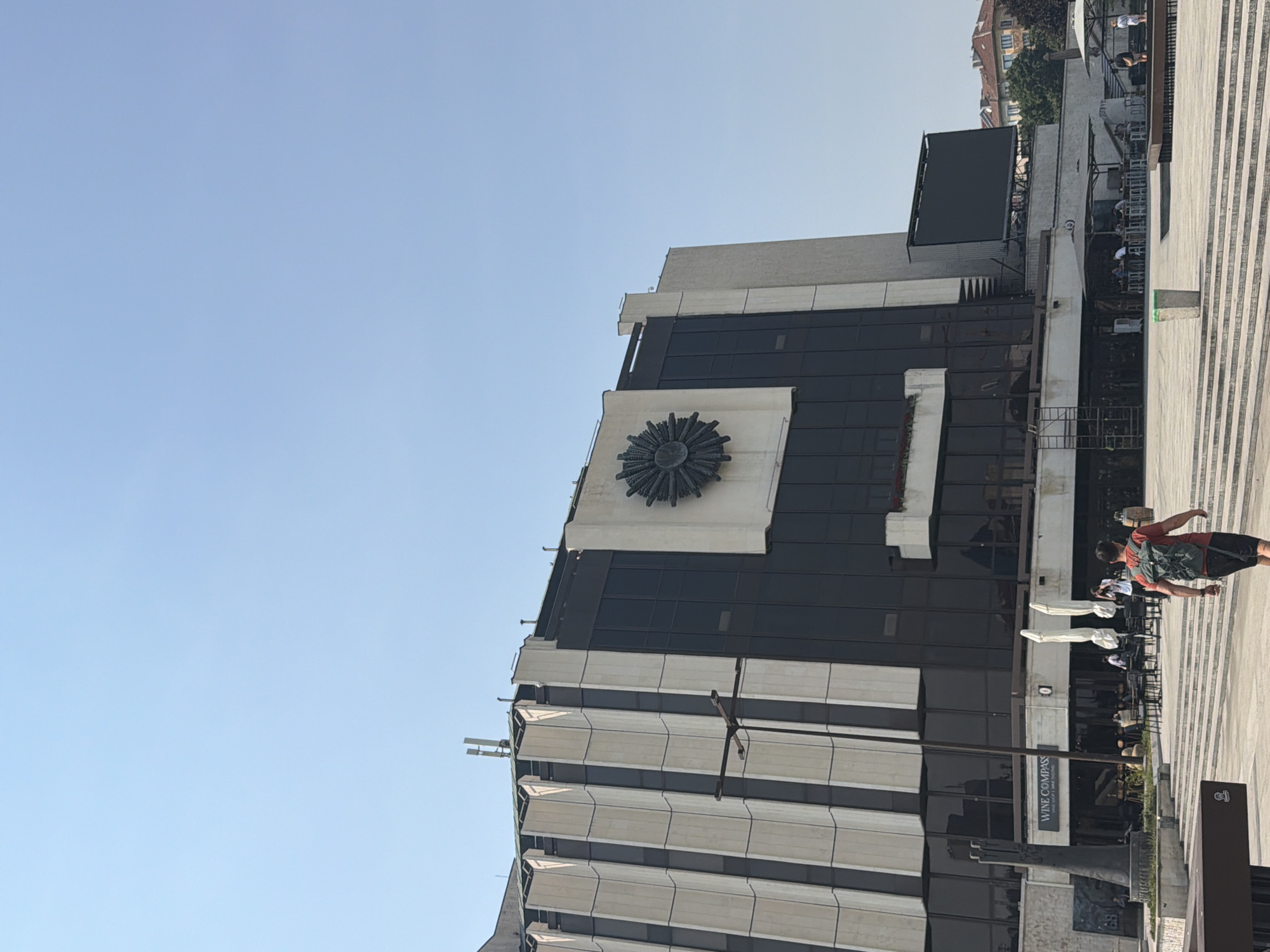 This media file captures a sunlit day in Sofia, Bulgaria, with a clear, light blue sky. The primary subject is a modern building with a dark, reflective facade composed of many windows. A prominent, sun-like circular sculpture is affixed to the building. The lower part of the building features a striped, light-colored facade. In the foreground, a person with a backpack is walking on a paved area with diagonal stripes. Several tables and chairs are visible in front of the building, suggesting an outdoor seating area. The overall scene is one of urban activity.
A sign visible on the building reads "WINE COMPASS". The presence of people suggests a public space during daytime hours, likely a cultural or commercial center. The lighting indicates it's a bright, sunny day, possibly late morning or early afternoon. The architecture and the overall setting are characteristic of a modern European city.