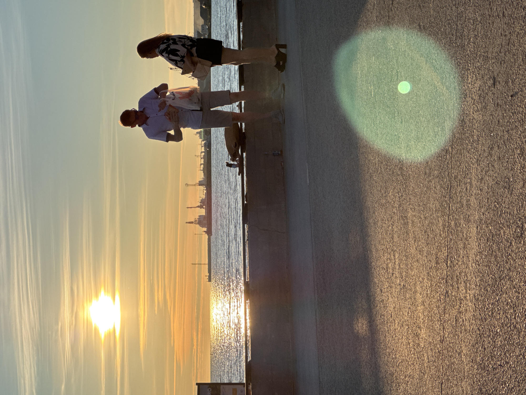 At sunset in Tallinn, Estonia, two individuals stand on a concrete promenade overlooking the sea. The sun, a bright orb in a sky streaked with wispy clouds, casts a golden glow on the water and the surrounding landscape. In the distance, ships and industrial cranes are visible, suggesting a port area. A man in a light-colored short-sleeved shirt and shorts holds a white plastic bag, and a woman in a dark printed top and skirt stands beside him. They appear to be engaged in conversation or simply enjoying the view. The promenade is paved with asphalt, and a concrete barrier separates it from the water. A green lens flare is prominent in the foreground, indicating the direction of the sun.