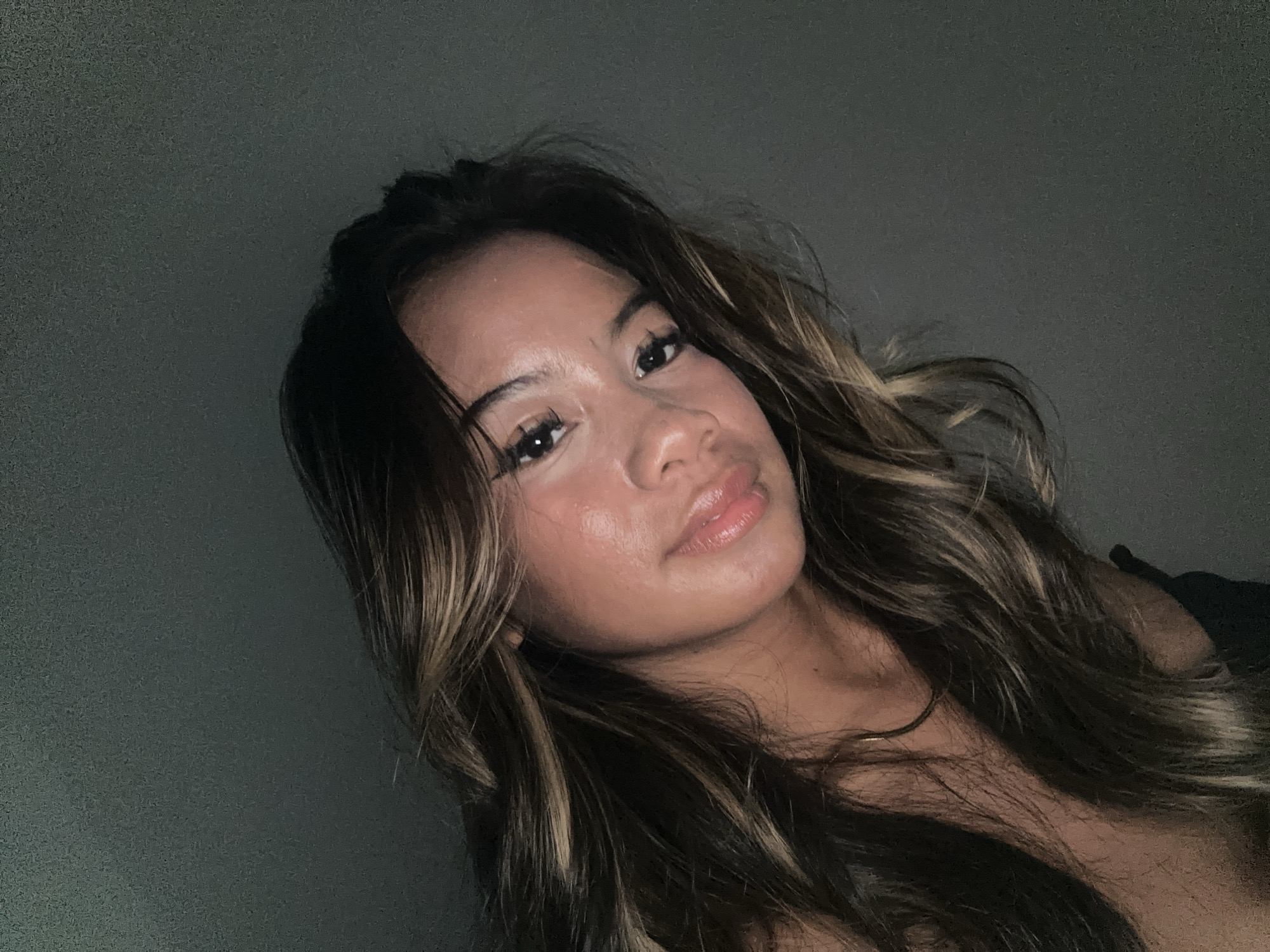 A medium-shot portrait of a young woman with shoulder-length, wavy, dark brown hair with blonde highlights. She is fair-skinned with full lips and dark eyelashes. The background is a plain dark grey wall. No city or specific location is discernible from the image.