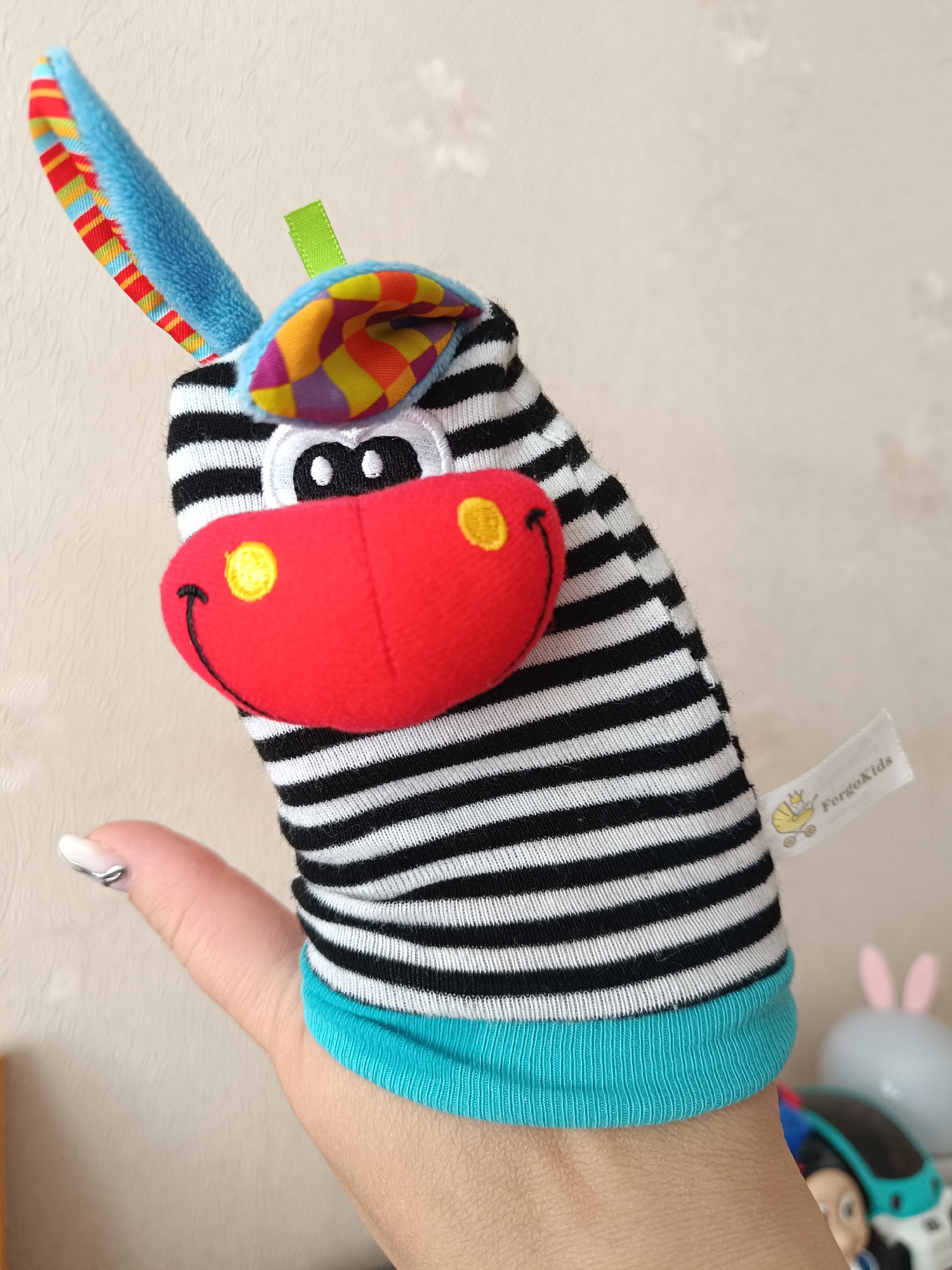 A hand puppet of a zebra is being held in someone's hand. The zebra puppet is black and white striped with a blue collar. It has a red nose and multicolored ears. The puppet has a tag on its side that reads "FergoKids". The background is a plain beige wall. The location is unknown and cannot be assumed to be Tobolsk, Russia based on the image.