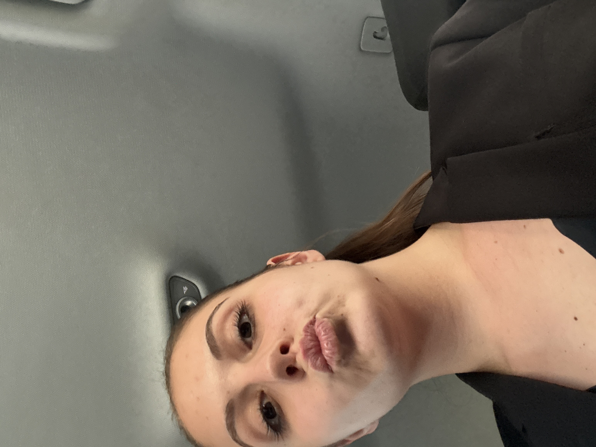 A young woman is the central subject of this close-up photograph, taken from a low angle within the interior of a vehicle. Her head is tilted, and her face, the primary focus, occupies the lower right portion of the frame. She has long, dark brown hair that cascades over her left shoulder and is also visible beside her face. Her eyebrows are dark and well-defined, matching her dark eyes, which are looking directly towards the camera. Her lips are pursed in a distinct "kissing face" or pout, conveying a playful or affectionate expression. She is wearing a dark, likely black, top or jacket, visible on her right shoulder and arm.
The background consists mainly of the light gray fabric of a car's headliner, suggesting an indoor setting within an automobile. In the upper-central part of the image, a small gray plastic hook or clip, commonly found in car interiors for hanging items, is visible. To the left of her ear, a small black plastic module, possibly a light fixture or control, is partially seen against the gray ceiling. The lighting is soft and even, without harsh shadows, consistent with natural light filtering into a car during daytime or subtle artificial interior lighting. There is no visible text in the image. The overall impression is a casual, intimate moment captured in a car. The location is Burgas, Bulgaria.