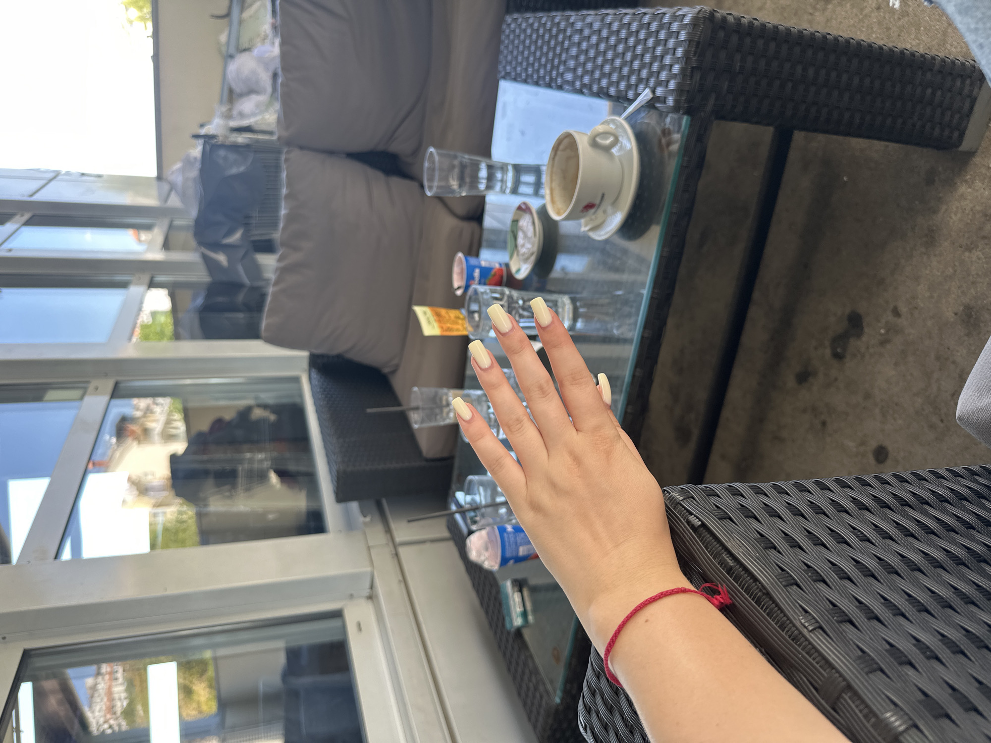 A person's left arm, adorned with a red string bracelet and light-colored fingernails, rests on the armrest of a dark, woven outdoor chair at a cafe. The arm is positioned in the foreground, leading the eye towards a glass-topped coffee table. On the table, several objects are visible: a white cup and saucer containing remnants of coffee, a tall glass of water, another glass with ice, a small container with a strawberry image, a pack of cigarettes, and a yellow card. In the background, a grey outdoor sofa with cushions is partially visible, alongside large windows reflecting buildings and sky. The cafe appears to be located in an outdoor seating area, possibly on a balcony or terrace, with concrete flooring. The setting suggests a casual daytime scene in Grad Rijeka, Croatia.