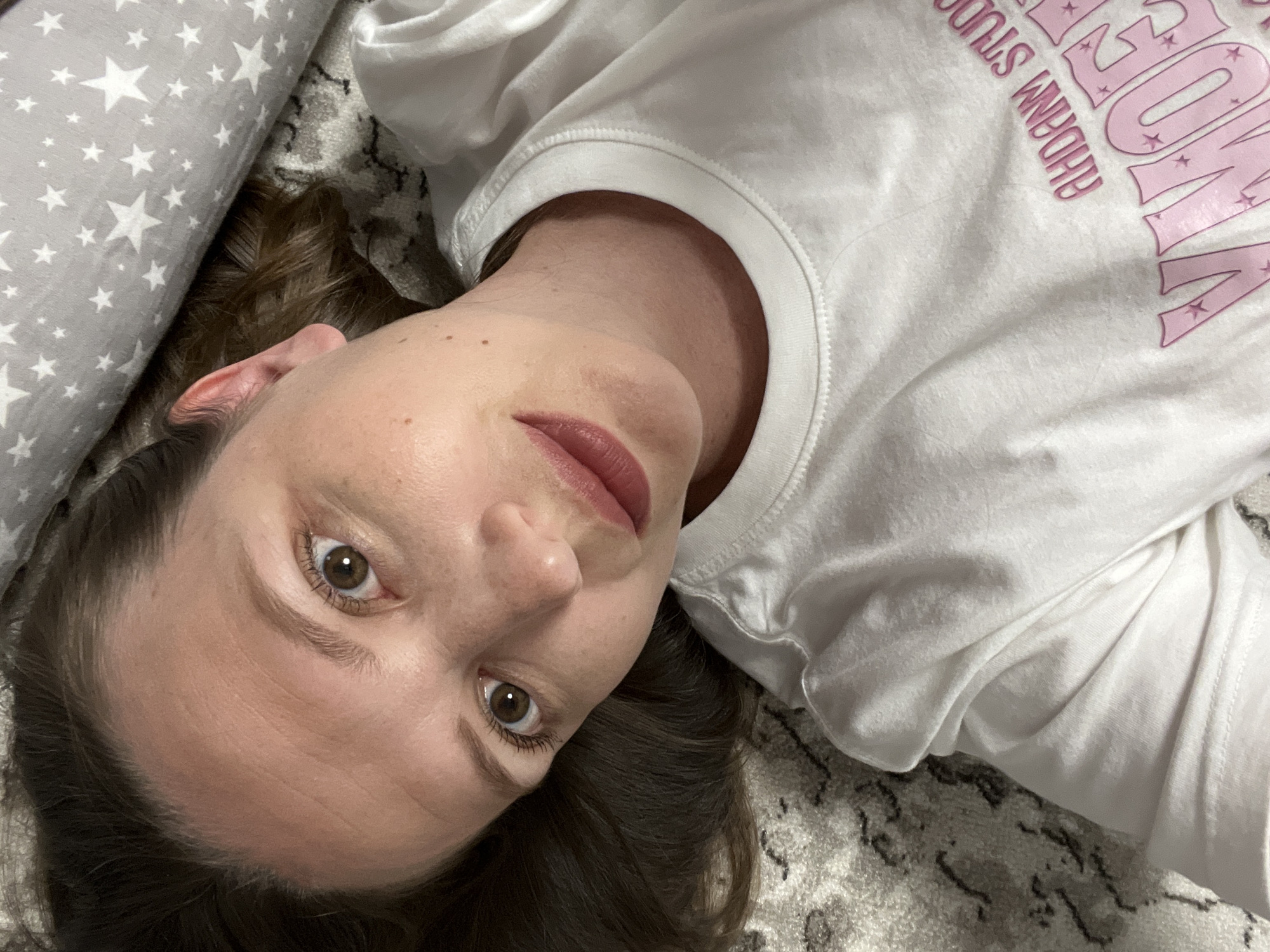A woman is lying on a patterned gray and white surface, possibly a carpet or rug. She is wearing a white t-shirt with pink lettering and lies with her head turned towards the camera. To her left is a gray pillow with white star patterns. The woman has brown hair, fair skin, brown eyes, and is wearing lipstick. The location information is not discernable from the image.