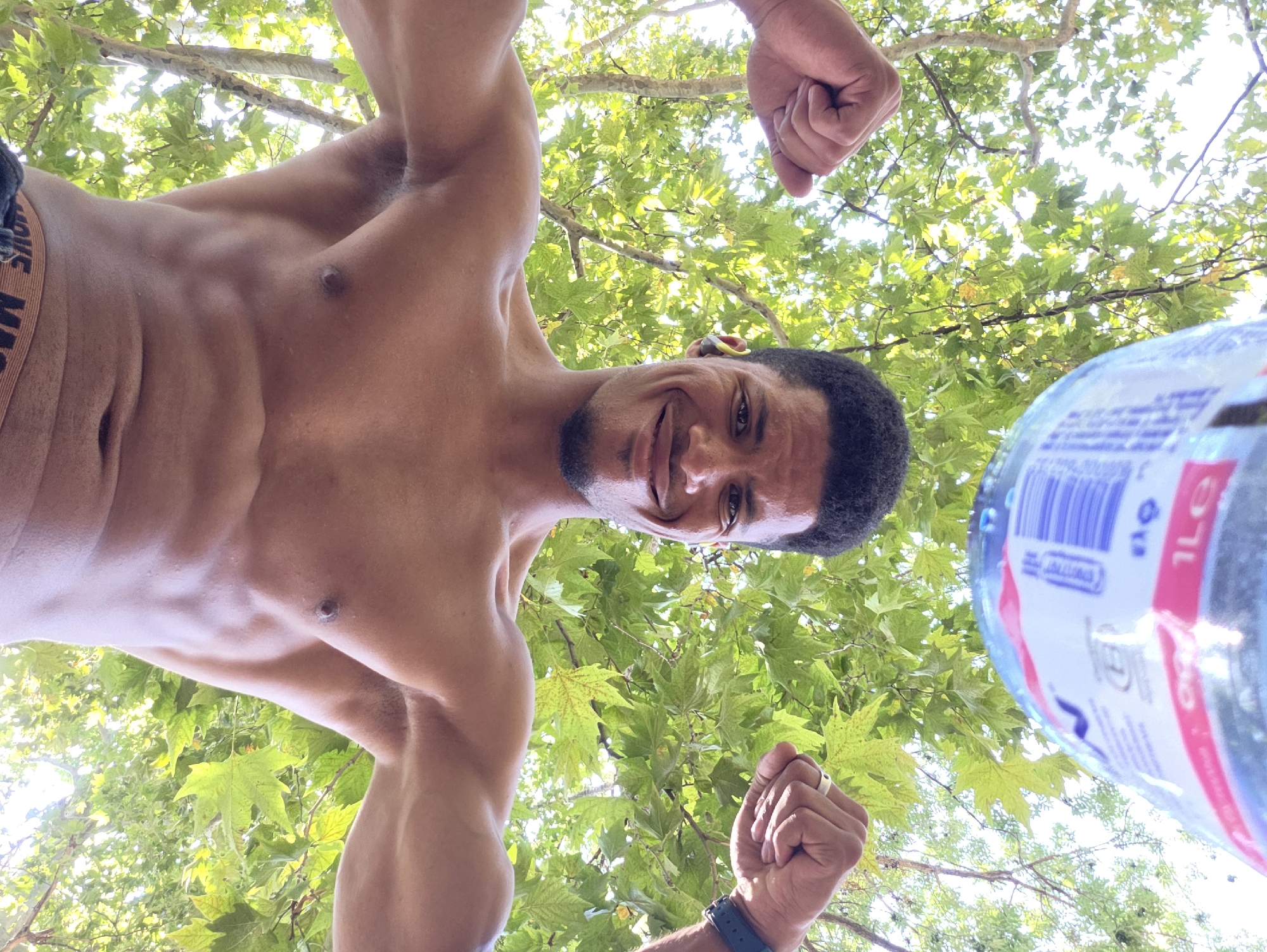 A low-angle, upward-facing shot shows a shirtless man with muscular arms and defined abs, flexing his biceps. He is smiling and looking directly at the camera. He has short, dark, curly hair and is wearing small, yellow earbuds. A large, clear plastic water bottle with a blue and white label is visible at the top of the frame, appearing to be held above him. The background consists of lush green leaves and branches of a tree, suggesting an outdoor setting. Sunlight filters through the leaves, creating bright highlights. The man is wearing patterned underwear. The image was captured in the city of Varna, Bulgaria.
