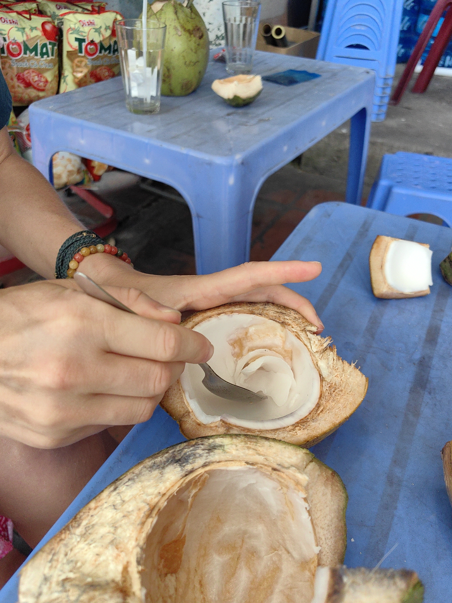 A close-up shot shows a person scooping the inside of a coconut with a spoon on a blue plastic table. Another half of the coconut sits below, already scraped clean. A piece of the coconut and its pulp is on the table next to it. In the background, on another blue table, are bags of Oishi brand snack food, a whole coconut, and a glass. Blue plastic chairs surround the tables. The location is Cao Xanh Ward, Vietnam.
