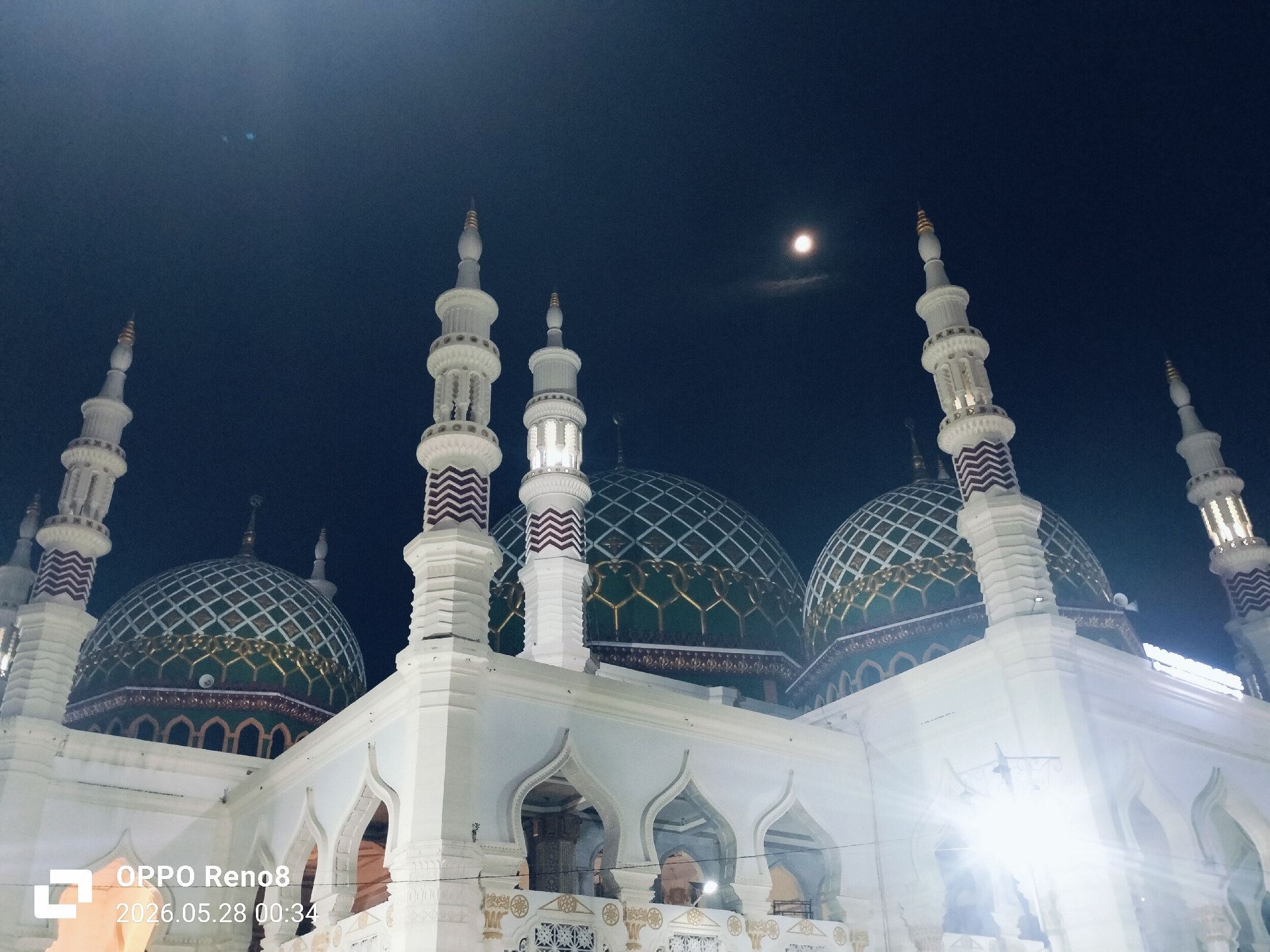 The image shows the Baitul Muttaqin Mosque in Kotapantonlabu, Indonesia, taken at night. The mosque features multiple green and gold domes with white latticework. The white exterior of the mosque is illuminated by strong artificial lights, creating a bright contrast against the dark night sky. Several tall, slender minarets with white and maroon accents rise from the mosque's structure. In the dark blue sky, a small, round light suggests the presence of the moon. At the bottom left corner of the image, there is a watermark indicating "OPPO Reno8" and a timestamp "2026.05.28 00:34". The perspective is from a low angle, looking upwards towards the mosque and the sky.
