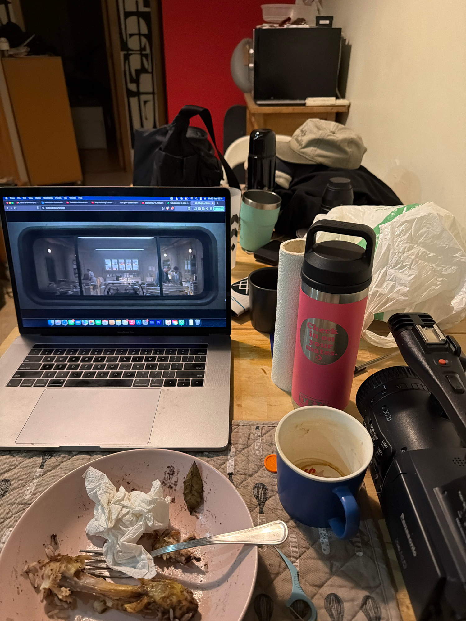 The image depicts a cluttered desk in what appears to be a room, possibly a home office or living space. The scene is viewed from a top-down perspective. On the left side of the frame is an open laptop displaying a scene from a television show or movie, showing people in what looks like a cafeteria or dining area. To the right of the laptop, there are several objects scattered across the desk: a pink water bottle with text that reads "Check in bit your mates.", a roll of paper towels, a blue mug with remnants of a beverage, a black video camera, a crumpled paper napkin on a pink plate with remnants of food (a bone and rice), and a fork. There are also other items visible in the background, including a black bag, a beige baseball cap, and a stack of plastic bags. The lighting suggests it is nighttime or a dimly lit room. The presence of a laptop, camera, and various personal items indicates a casual, possibly work-related or leisure setting. There is no indication of the specific location within the city of Almada, Portugal.