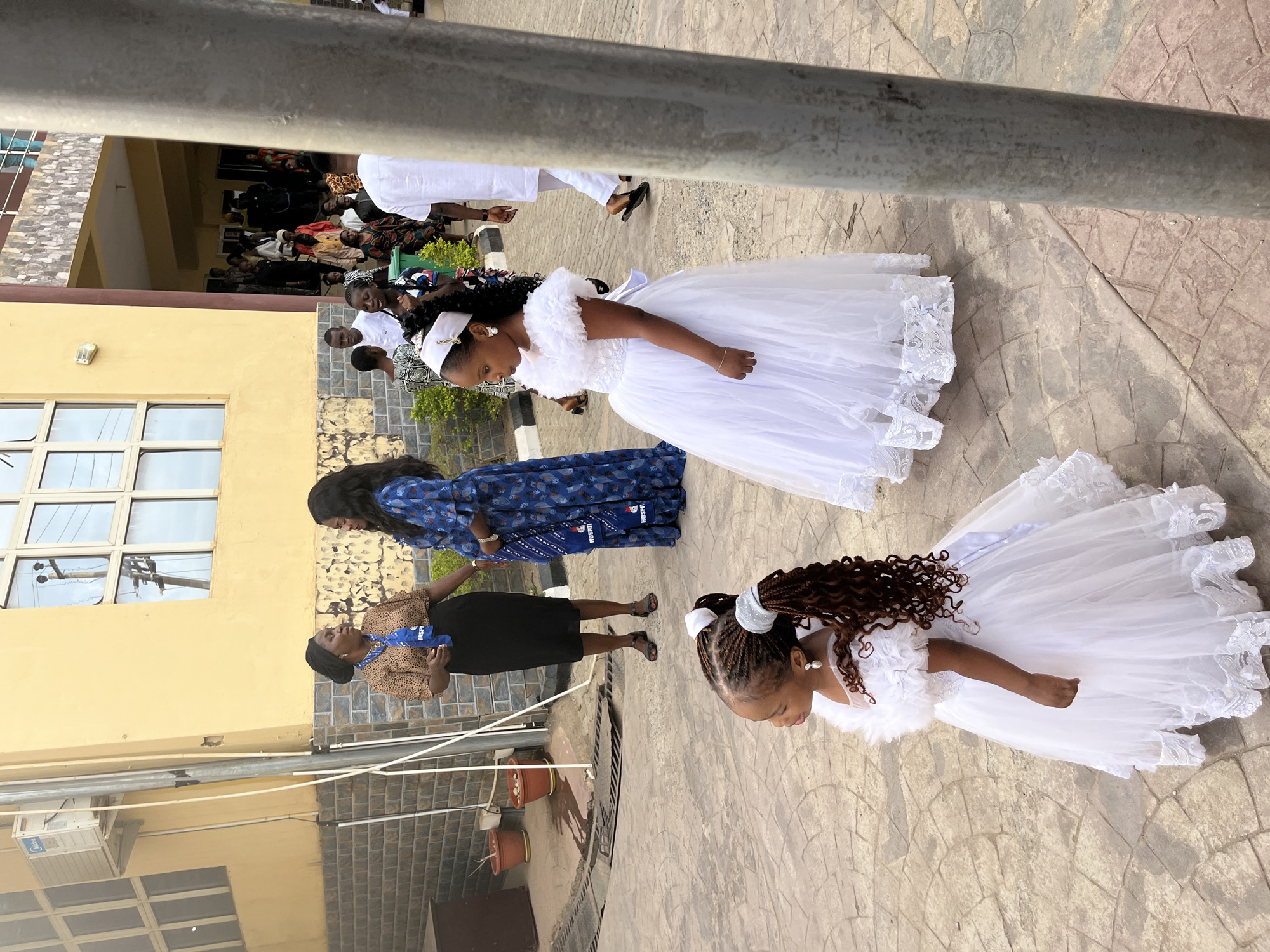 The image shows two young girls in white dresses, likely flower girls, standing on a paved outdoor area in what appears to be Abuja, Nigeria, based on the people's attire and skin tones. One girl has brown hair and a white headband, while the other has black hair and a white headband. Two women stand behind them, one in a blue patterned dress and the other in a brown patterned top with a black skirt and blue lanyard. A group of people is visible in the background near a building with yellow walls and barred windows. Potted plants are on the ground. A metal pole is visible at the top of the frame.