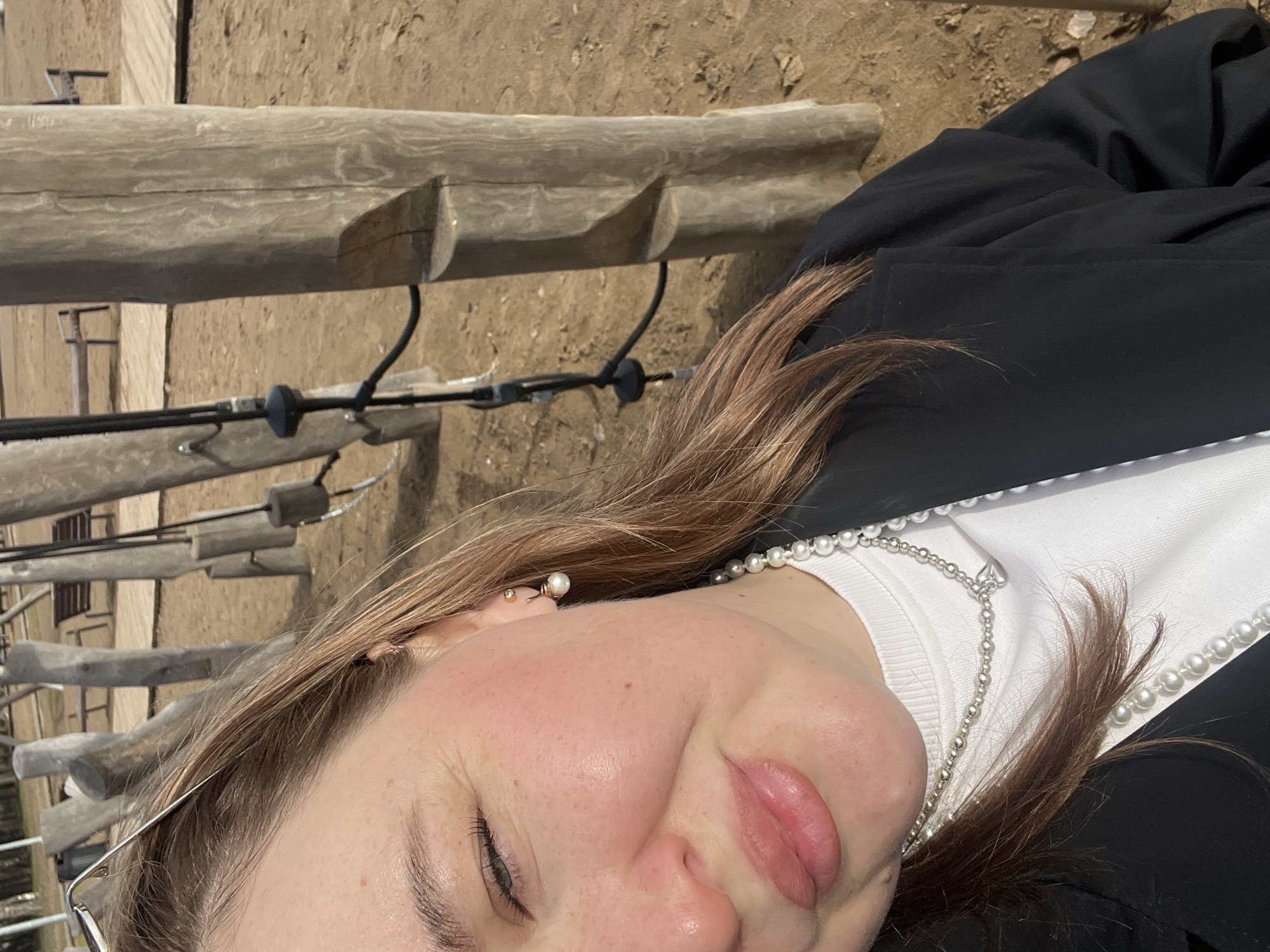 A close-up, slightly angled, daytime shot features a person's face, primarily the right side, alongside an outdoor setting with weathered wooden structures. The person, likely female, exhibits fair skin, with prominent cheek coloration, and a single visible eye with light-colored lashes. Her hair is light brown, parted, and partially covers her clothing. She is wearing a white top with pearl embellishments around the neckline, partially covered by a black jacket. A silver chain and a stud earring are visible.
In the background, weathered, pale wooden beams run horizontally. Beneath these beams, black cables are suspended by hook-like structures. The ground is composed of dirt, and other architectural elements are faintly visible in the distance. The location is presumed to be in the city of Chaikovsky, Russia.