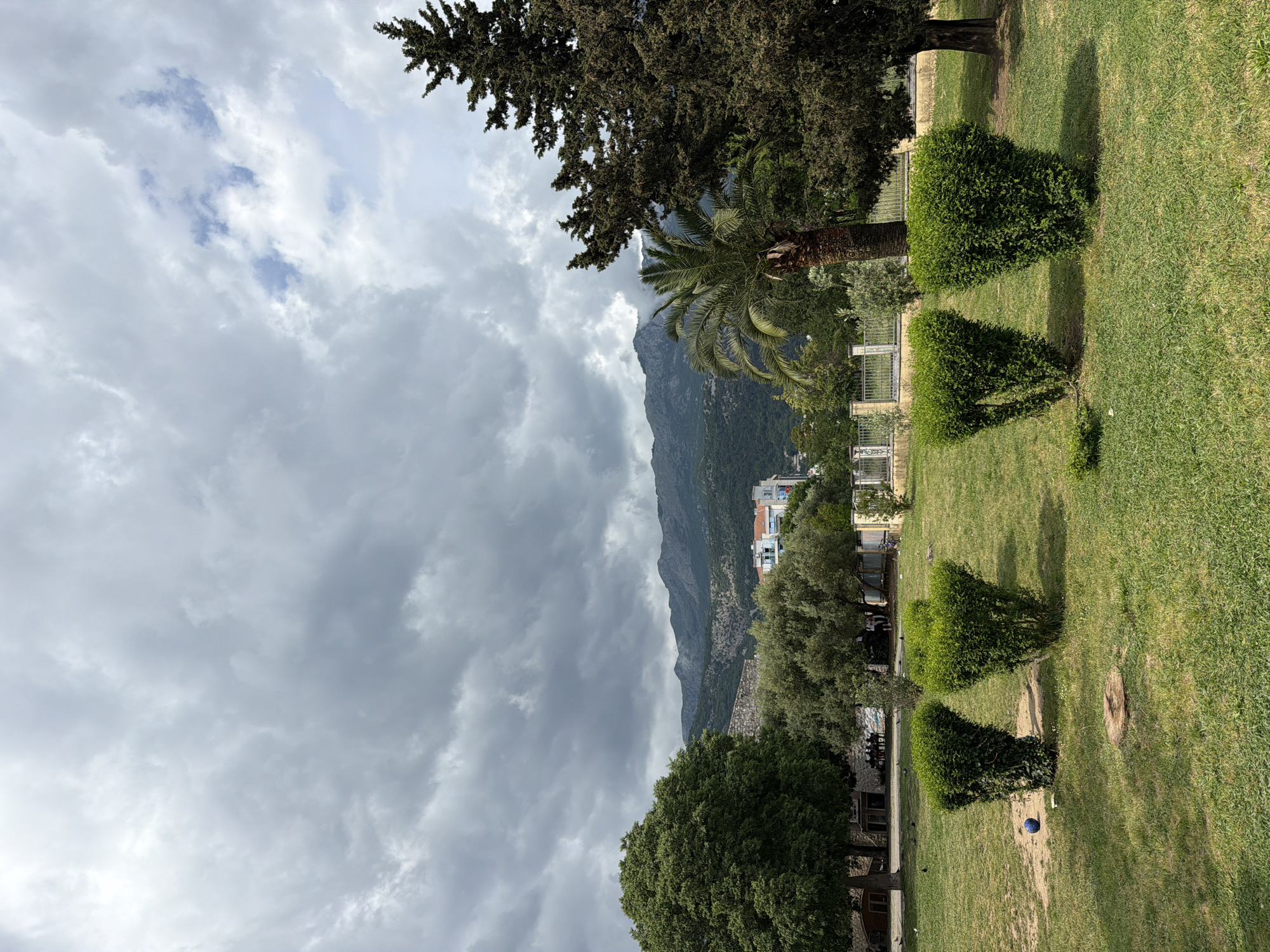A landscape photograph captures a park scene in Budva, Montenegro, under a cloudy sky. In the foreground, the park's grassy lawn is dotted with meticulously trimmed hedges. A line of trees and a metal fence separates the park from the urban area. The background features a towering mountain partially obscured by clouds, with buildings and houses nestled on its lower slopes. A palm tree stands amidst other trees, adding to the Mediterranean feel.