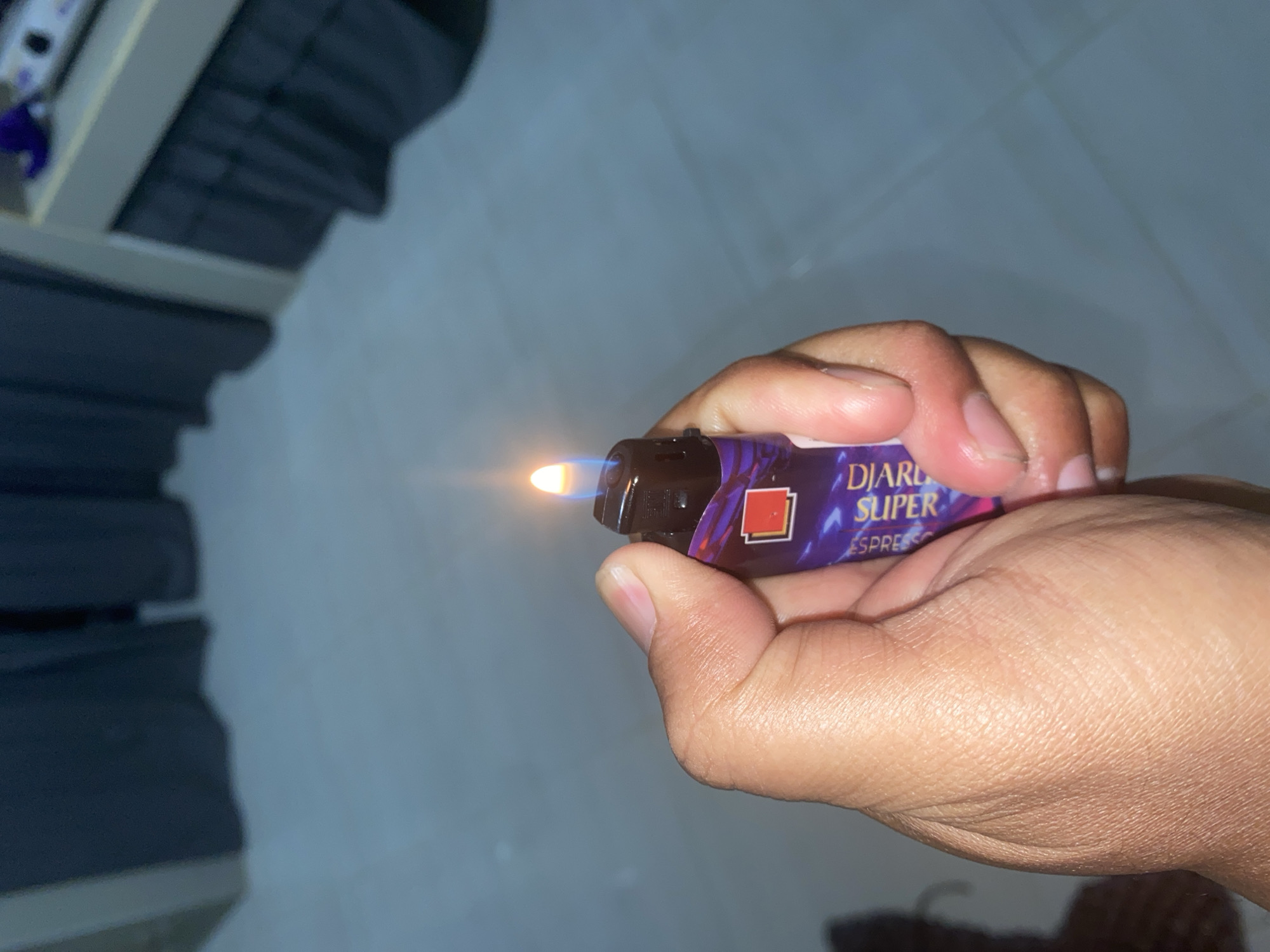 A close-up shot captures a hand holding a lit lighter with a purple and blue design, displaying the text "DIARIO SUPER ESPR." The flame is visible at the top of the lighter, emitting a warm glow. The background is blurred, showing dark curtains and a light-colored floor, suggesting an indoor setting. The image is not geotagged and therefore cannot be definitively located in Bontoharu, Indonesia.