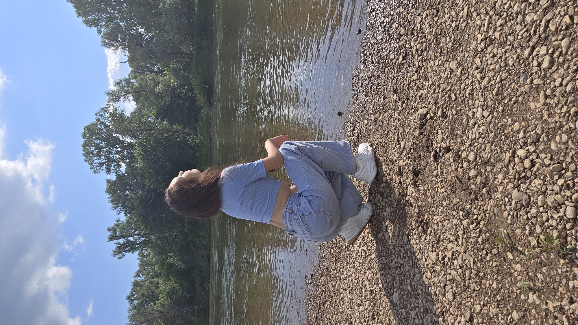 A young woman with brown hair is squatting on a rocky shore next to a river. She is wearing a light blue crop top and light blue jeans, with white sneakers. The river is calm and reflects the blue sky with scattered white clouds. The opposite bank of the river is covered with lush green trees. The sun is shining, casting shadows on the rocky ground. The scene appears to be in a natural setting, likely in or near the city of Darino, Russia, given the vegetation and river characteristics.