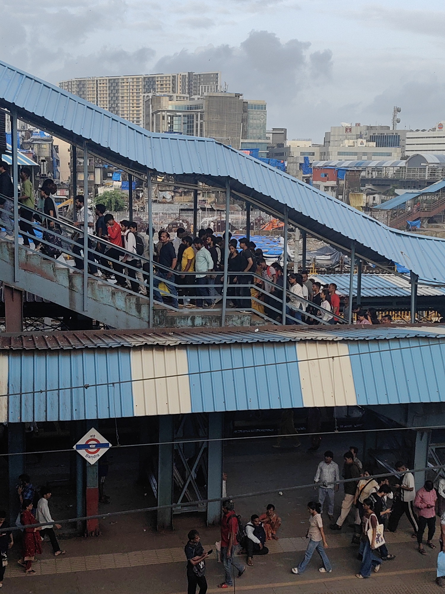 This image captures a bustling scene at the Bandra railway station in Mumbai, India. A crowded pedestrian overpass, constructed with blue corrugated metal roofing and supported by metal beams, dominates the upper portion of the frame. Numerous people are walking up and down the metallic stairs of the overpass, most appearing to be commuters. Below the overpass, the ground level of the station is visible, with more people milling about, some sitting, others walking. A diamond-shaped station sign with the text "Bandra" in English and possibly Hindi or Marathi is affixed to a blue and white striped awning. The background reveals a dense urban landscape with tall buildings, indicative of Mumbai's metropolitan environment. The sky is overcast, suggesting cloudy weather.
