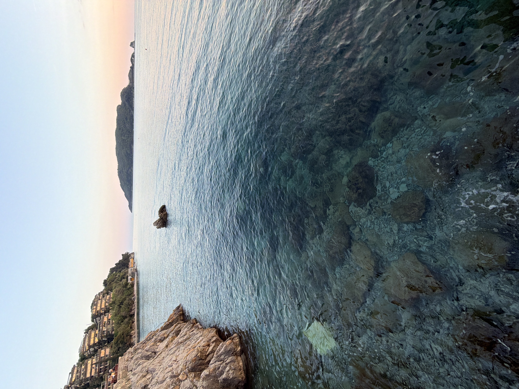 This image depicts a serene coastal landscape in Budva, Montenegro, likely captured during the golden hour of late afternoon or early evening. The scene is characterized by calm, clear waters and a rugged, inhabited coastline under a soft, gradient sky.
The right side of the frame features the sea, exhibiting remarkable transparency, especially near the shore. The clear water reveals a rocky seabed with various shades of grey, blue, and green stones, visible through the gently rippling surface. The water deepens further from the shore, transitioning from a light turquoise to a darker, more profound blue. A distinct, dark rock formation stands isolated in the water further out from the coast. In the bottom right foreground, a lighter-colored, possibly plastic, object floats near the rocky edge.
On the left side of the image, a verdant, tree-covered hillside slopes down to meet the sea. Numerous buildings, characterized by their architectural style with balconies and warm-toned facades, are nestled into the hillside, appearing to cascade towards the water. These structures reflect the soft, golden light of the low sun. In the distance, along the horizon, a larger, darker mountain range or landmass is silhouetted against the sky, defining the expansive backdrop. The sky above transitions from a clear, pale blue at the top to soft hues of pink and orange closer to the horizon.
The overall atmosphere is peaceful and tranquil, highlighting the natural beauty of the coastal environment. There is no visible activity or identifiable text within the image.