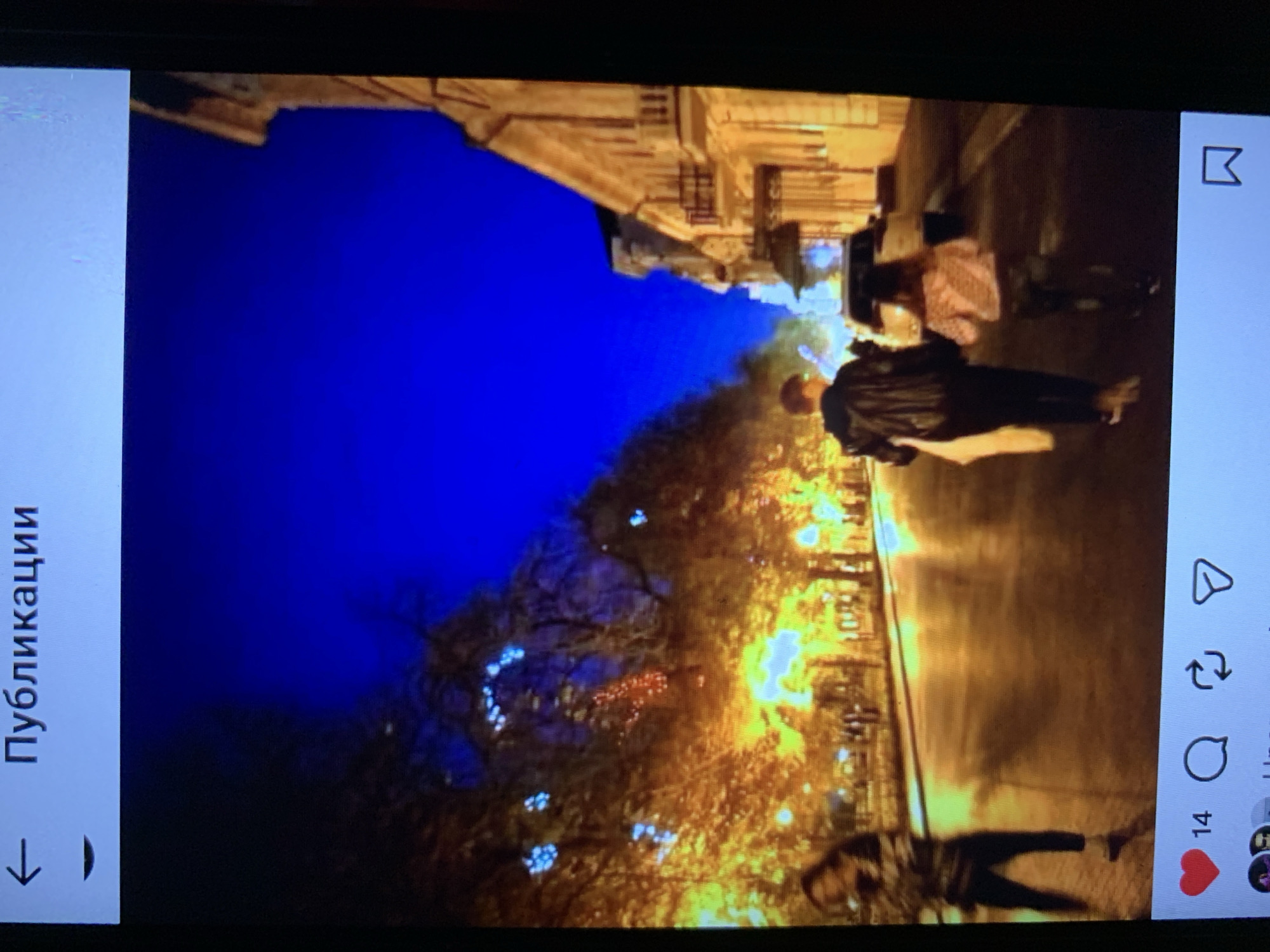 The image depicts an outdoor scene at night, illuminated by streetlights and decorative lights on trees. The sky is a deep, vibrant blue. On the left side, a woman in dark clothing and leggings is walking away from the camera, her back to the viewer. Further down the street, a couple, a man and a young girl, are walking hand-in-hand away from the camera towards a white car. The man is wearing a dark jacket and pants, and the girl is wearing a patterned jacket and dark pants. The buildings on the right side of the street are illuminated, showing architectural details. The street itself is wet and reflects the lights. The foreground of the image shows a social media interface with icons for likes, comments, shares, and saves, as well as a number "14" indicating likes. The location is identified as Lymanka, Ukraine.