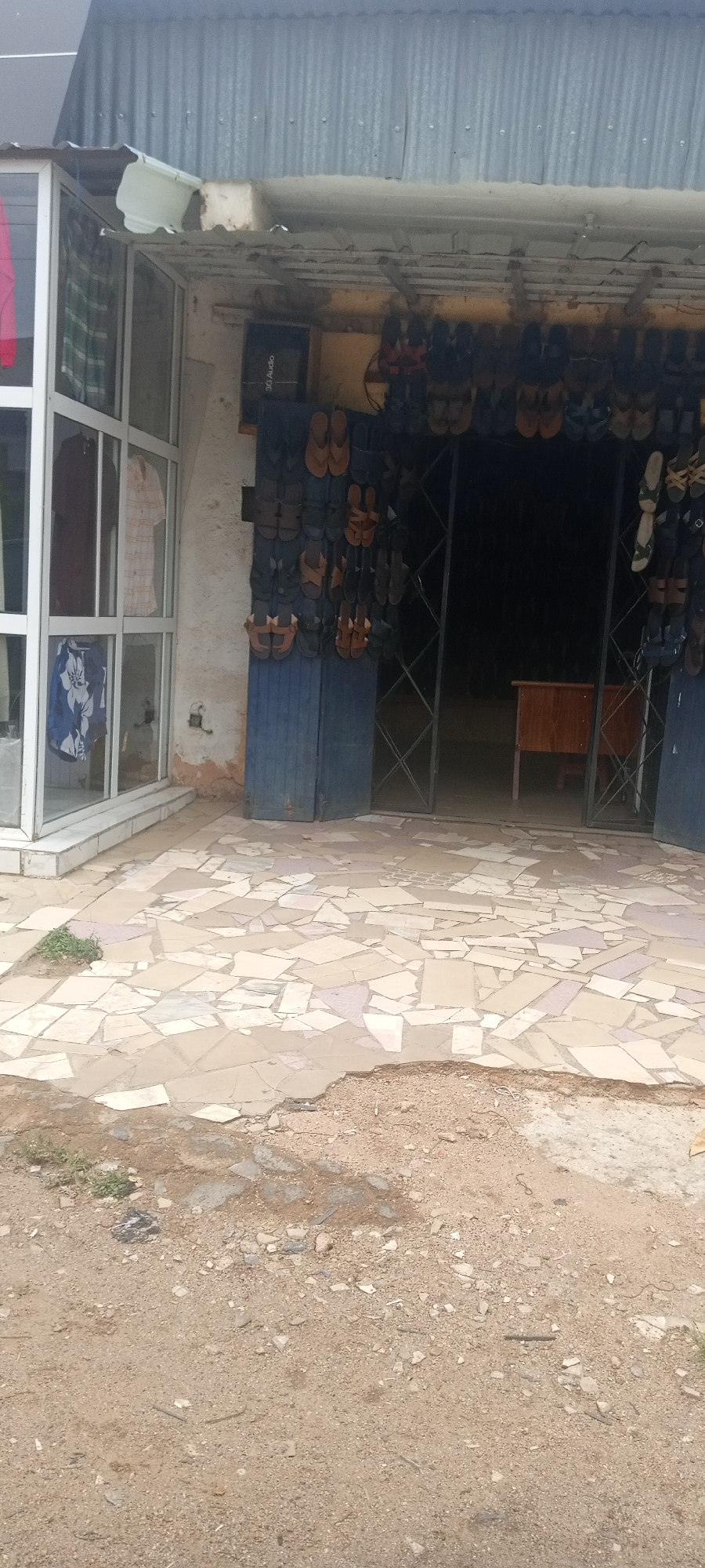 The image shows a shop in Jos, Nigeria, selling sandals and clothing. The shop entrance is lined with hanging sandals. A clothing display is visible through a glass panel to the left. The ground in front of the shop is a mix of cracked tile and dirt. The shop appears to be in a relatively modest or less affluent area. There are no people clearly visible in the image.