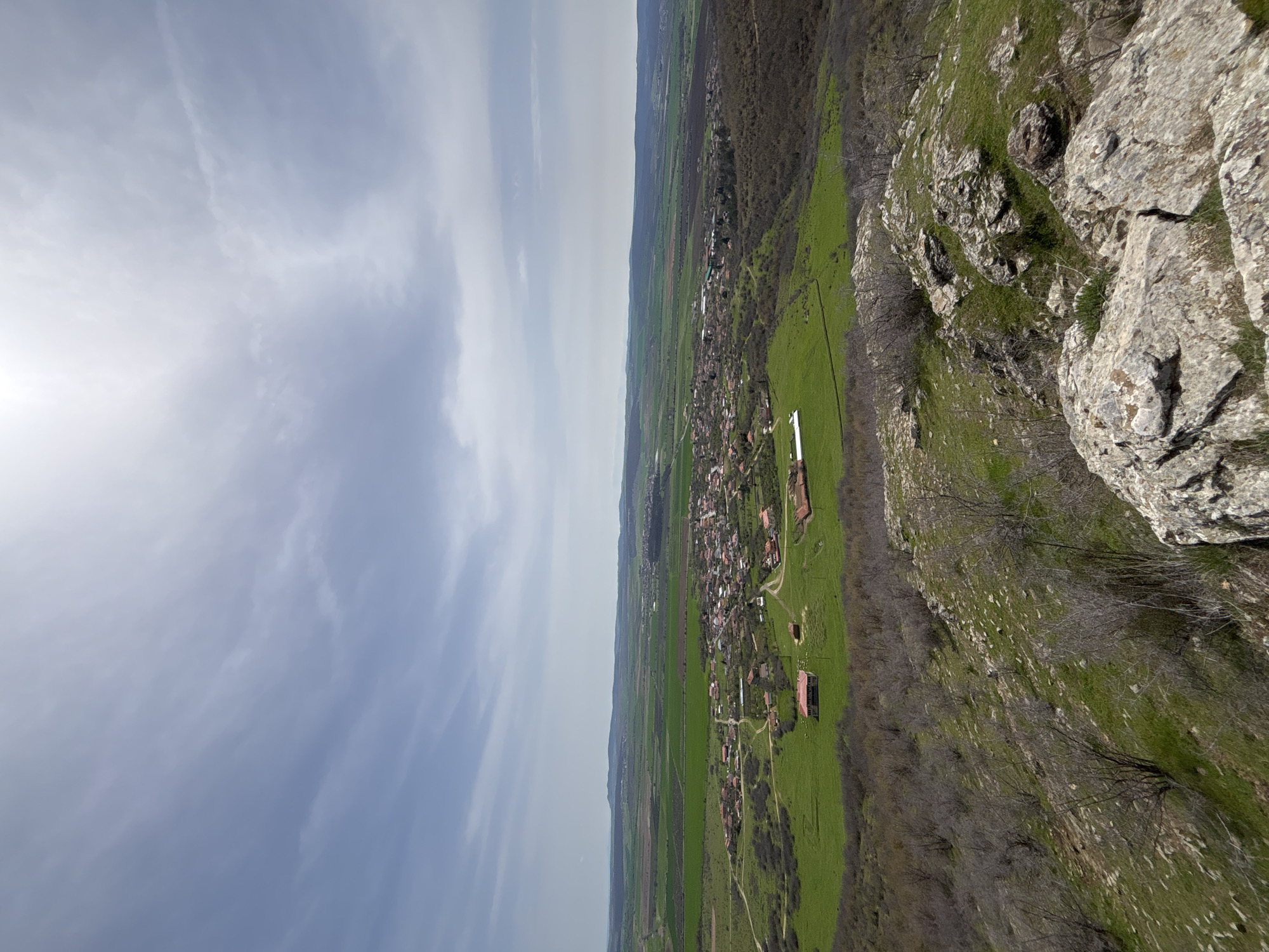 The image captures a landscape view in Goritsa, Bulgaria, showcasing a blend of natural and human elements. On the right side, a rocky terrain with sparse vegetation leads down to a green, grassy area interspersed with trees. A small settlement with buildings, many with red roofs, is visible along a central stretch, appearing as a linear progression across the green landscape. Beyond the settlement are fields of agricultural land. The sky is predominantly cloudy, with a light blue hue and hints of sunshine on the left side. The perspective is from an elevated position, giving a broad overview of the terrain.