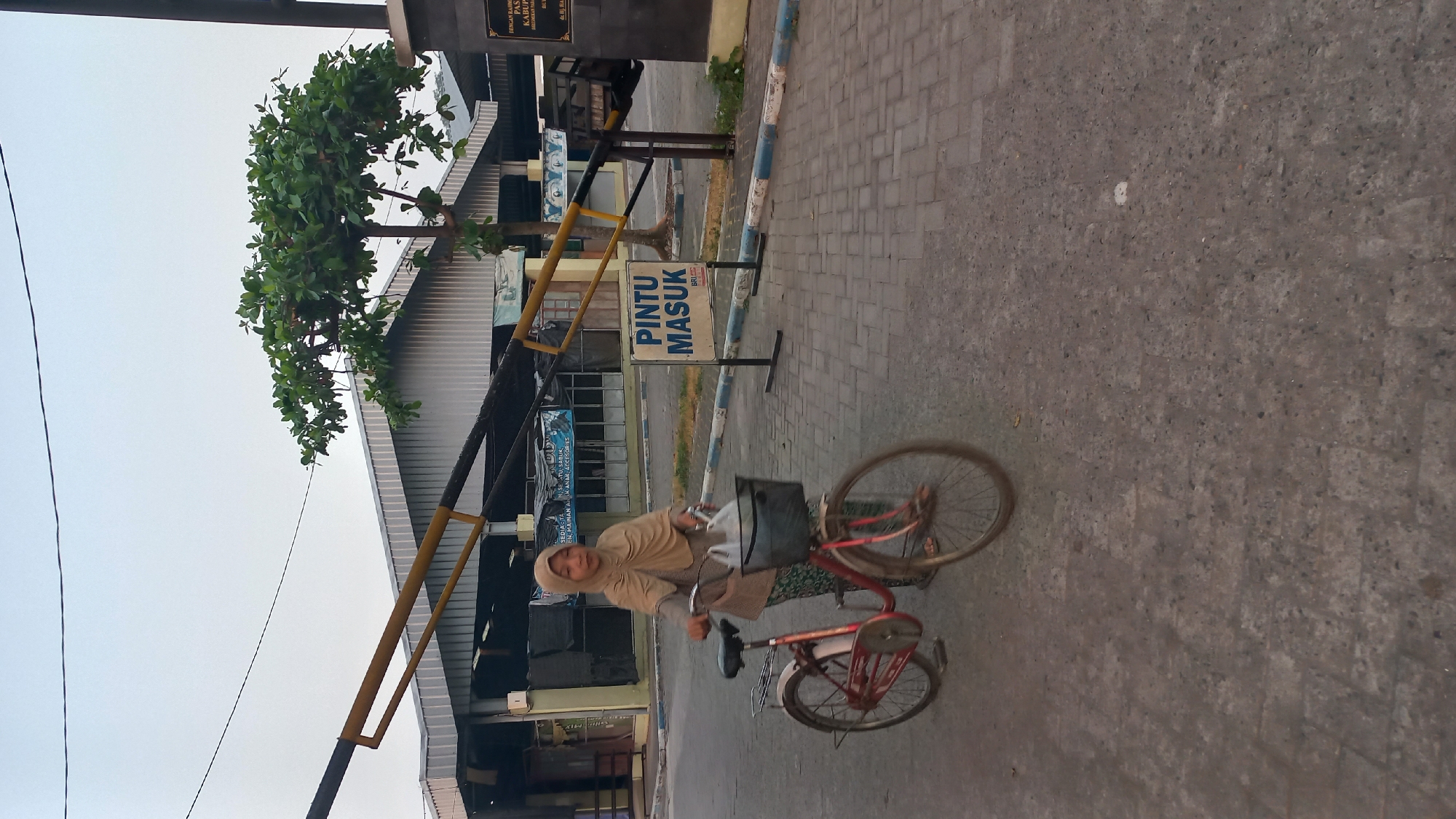 A young girl wearing a hijab rides a bicycle with a basket in front. She is dressed in a long-sleeved shirt and patterned pants. The bicycle is red with a silver basket holding plastic bags. The girl appears to be on a paved walkway with brick patterns, which leads to a gate. A sign nearby reads "PINTU MASUK," meaning "ENTRANCE." In the background, there is a large building with a metal roof, and a tree with green leaves. The sky is overcast. The image is likely taken in Pare, Indonesia, given the sign in Indonesian.