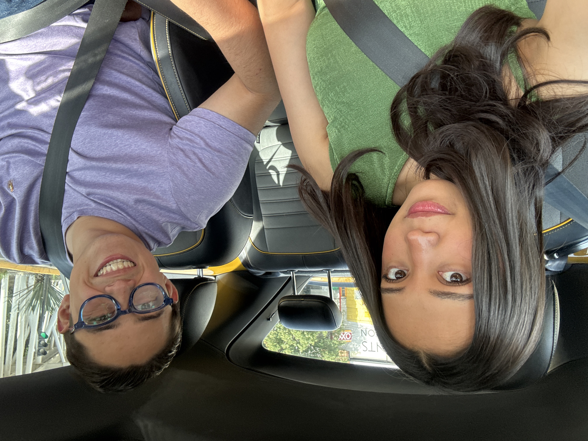 Two people, a male and a female, are visible inside a car. The man is wearing a purple shirt and blue-framed glasses, and he is smiling. The woman has long, dark hair and is wearing a green shirt. Both are wearing seatbelts. The interior of the car features black leather seats with yellow accents. Outside the car window, a sign is visible with the words "RENTS ON" and "OX" indicating "OXO". The location is Puebla, Mexico.
