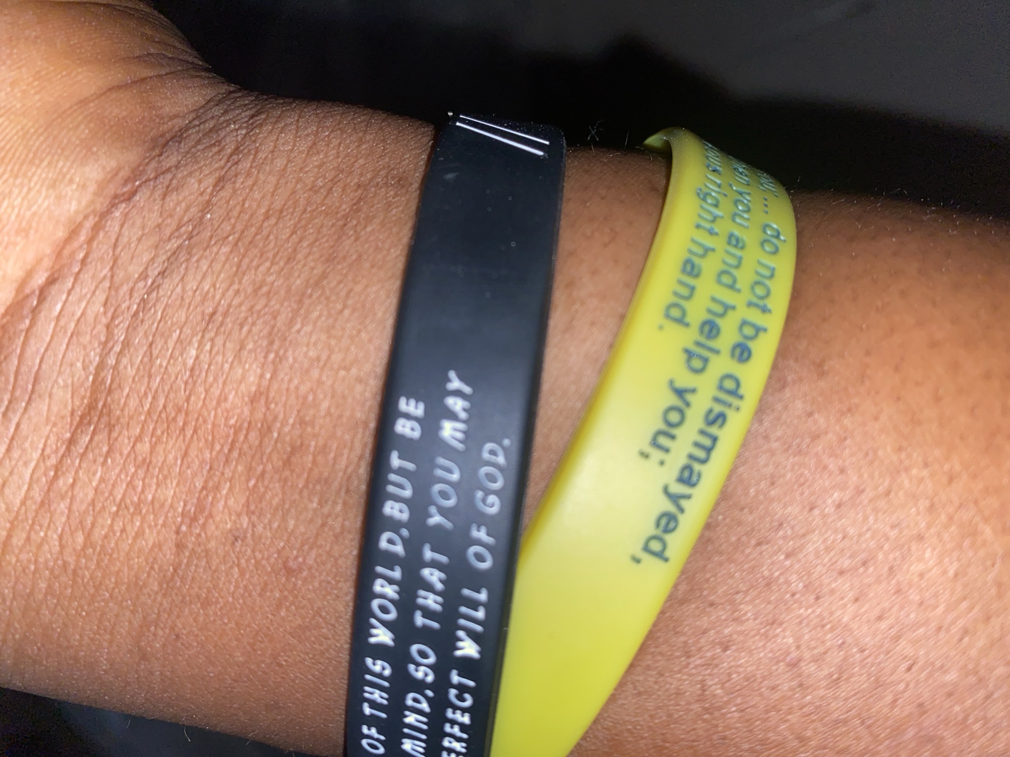 This is a close-up, indoor shot of a person's forearm wearing two silicone wristbands. The wristbands are black and yellow. The black wristband has white text that reads "OF THIS WORLD. BUT BE MIND, SO THAT YOU MAY ERFECT WILL OF GOD." The yellow wristband has text in blue that is partially visible and appears to read "... you and help you: do not be dismayed, ...vous right hand." The wrist is brown, and skin texture is visible. The background is dark and out of focus. There is no visible indication of location in Jauro Yinu, Nigeria.