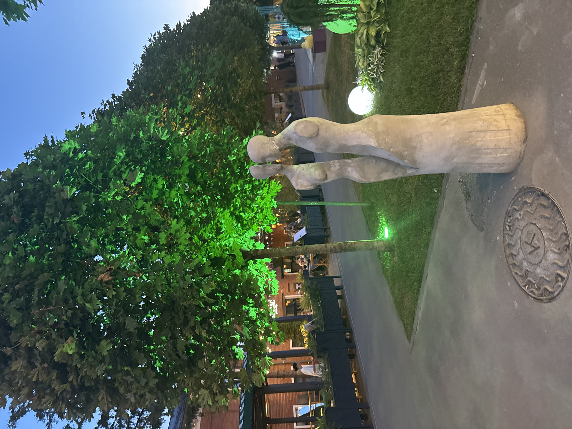 In Tula, Russia, an outdoor dining area is depicted at dusk. The scene features several trees illuminated with bright green lights, casting an artificial glow on their leaves and the surrounding environment. In the foreground, two abstract concrete sculptures of human figures stand side-by-side. A pathway runs alongside the dining area, with a manhole cover visible on the ground in the lower left corner of the image. Further back, patrons are seated at tables under canopies, with brick buildings forming the backdrop. The overall ambiance suggests a contemporary urban setting with a focus on outdoor leisure and aesthetic lighting.