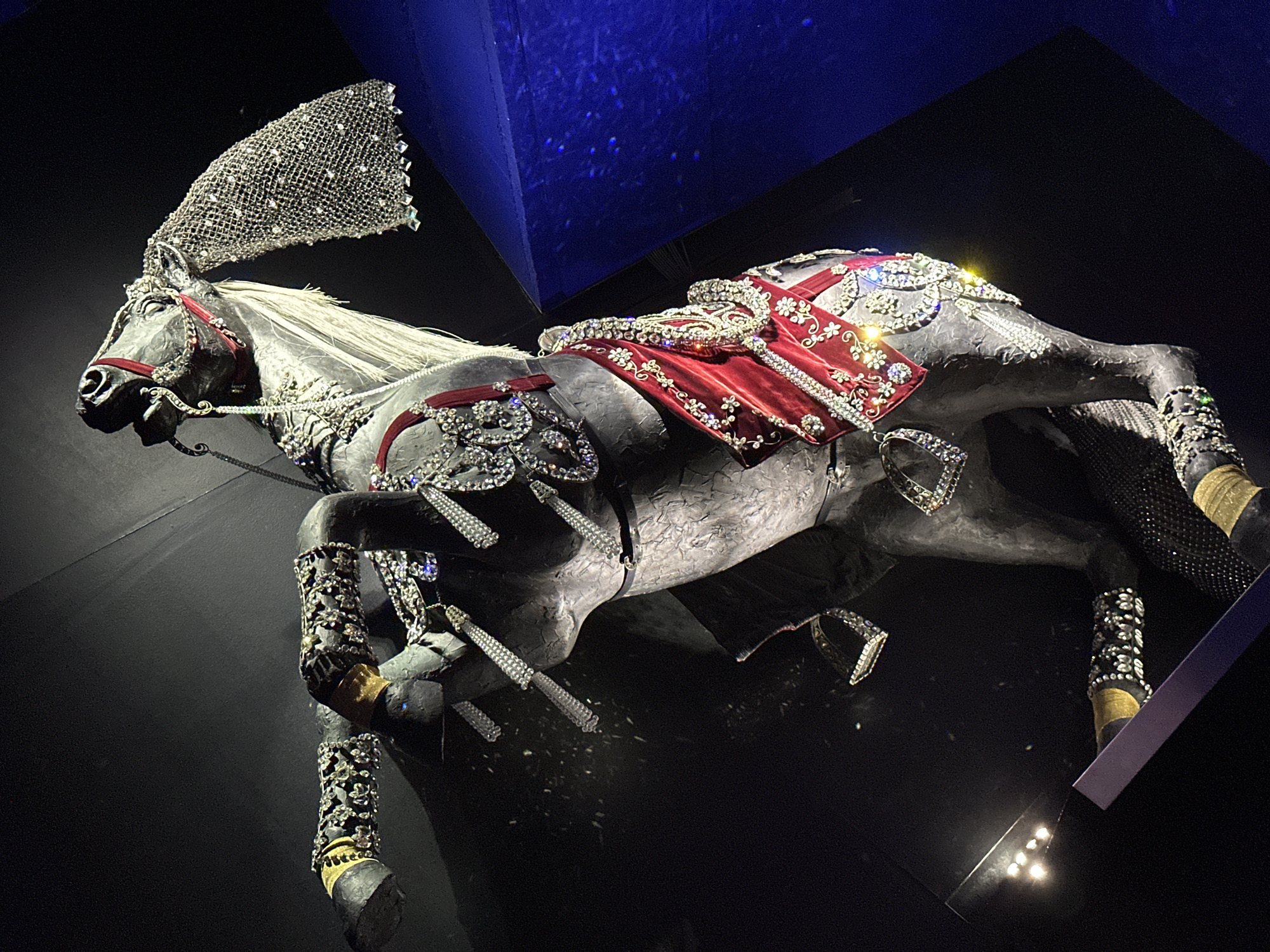 The image shows a highly ornate, life-sized sculpture of a horse, dramatically lit against a dark background with a blue, angular structure to the right. The horse is in a rearing pose, with its front legs raised. It is covered in a multitude of embellishments, including sparkling rhinestones and intricate silver detailing. The horse wears a red and gold draped saddle or cloth across its body, also heavily adorned with jewels. A mesh-like, crystal-encrusted veil or mane extension hangs from its head. The sculpture is on display in an exhibition space, likely within the Swarovski Kristallwelten (Crystal Worlds) in Wattens, Austria, given the context of elaborate crystal displays and the architectural style visible. The lighting is dramatic and theatrical, focusing attention on the detailed artistry of the horse sculpture.