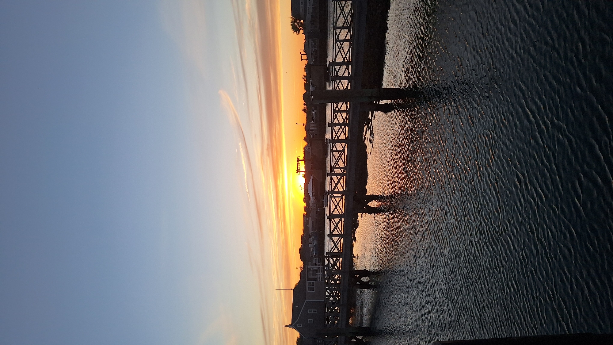 An outdoor, vertical, full shot captures a picturesque sunset over a waterfront town on Nantucket, USA. The sky transitions from a pale blue at the top to vibrant shades of orange and yellow near the horizon, with thin, wispy clouds streaking across the scene. A wooden pier extends from the lower left of the frame, crossing the dark, rippling water. The pier features a patterned railing with crisscrossed wood. On the left side of the pier, a gray building with a steeple stands. In the background, a line of buildings and trees is visible, silhouetted against the colorful sky. The setting sun casts a warm reflection on the water's surface.