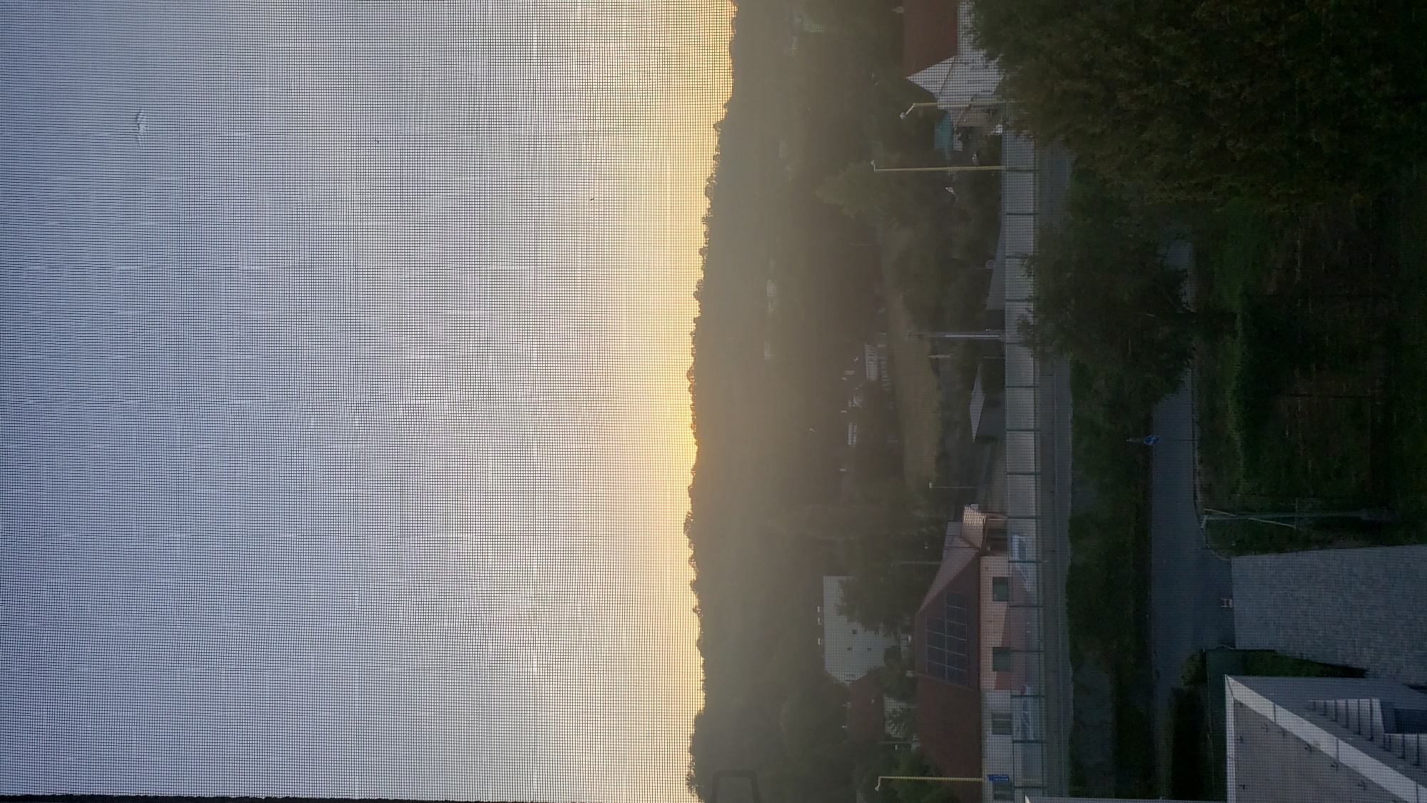 The image captures a hazy sunrise viewed through a window screen, with a silhouetted landscape of trees and buildings in Prešov, Slovakia. The sky transitions from a pale blue at the top to a warm yellow-orange near the horizon, indicating the sun is just rising or setting behind a hill. The foreground shows a glimpse of urban elements, including roofs of houses, solar panels on one roof, a street, and greenery. The overall impression is a tranquil, early morning or late evening scene in a residential area of the city.