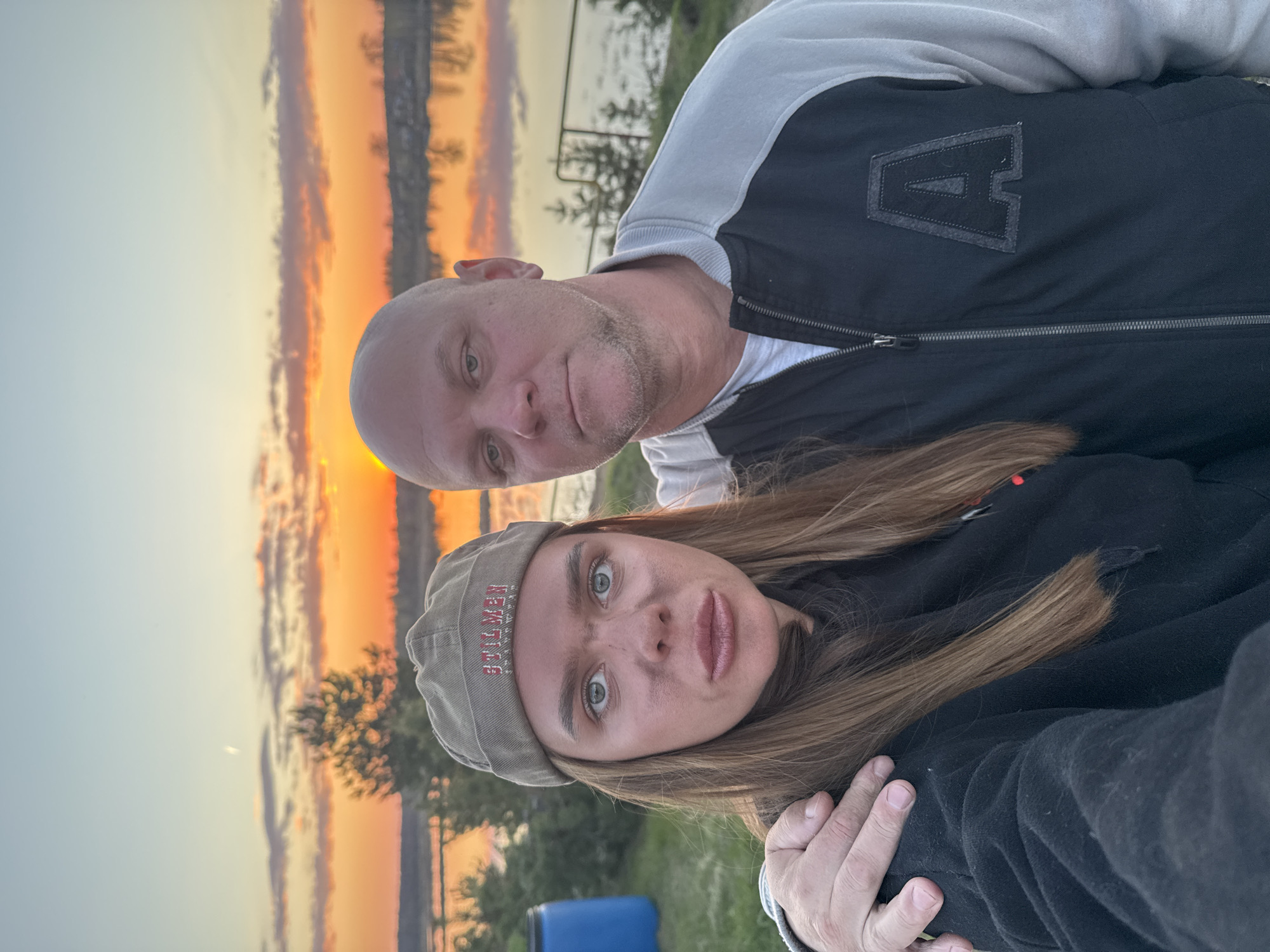 A selfie of two people with a sunset in the background. The man is fair-skinned, bald, and wears a jacket with a large "A" on the front. The woman has long brown hair and wears a cap. The location is identified by the river on the horizon, indicating it is near a body of water in Bol'shoye Kozino, Russia. The sky shows hues of orange and yellow indicative of a sunset.
