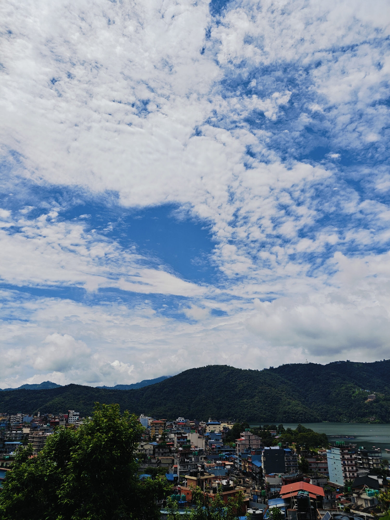 The image depicts an aerial view of the city of Pokhara, Nepal, with a focus on the Lakeside area. The foreground is dominated by lush green foliage, suggesting a high vantage point. Beyond the trees, a dense urban landscape unfolds, characterized by a multitude of multi-story buildings with varied architectural styles and colors, including blues, yellows, reds, and whites. Some buildings appear to be hotels or guesthouses, indicative of a tourist area.
In the mid-ground, a tranquil lake is visible, its surface reflecting the sky and the surrounding greenery. On the lake, several boats, including what appear to be traditional wooden boats and possibly some larger vessels, are present. The cityscape extends along the lake's edge, suggesting a vibrant lakeside community.
The background is characterized by rolling, densely forested hills that rise behind the city. These hills form a natural backdrop, with the vegetation appearing a deep, rich green. The sky above is a striking blue, interspersed with dramatic white clouds that create patterns of light and shadow across the landscape. The clouds vary in density, from wispy streaks to larger, more voluminous formations, contributing to the dynamic atmosphere of the scene. The overall impression is one of a bustling city nestled harmoniously within a beautiful natural environment.
