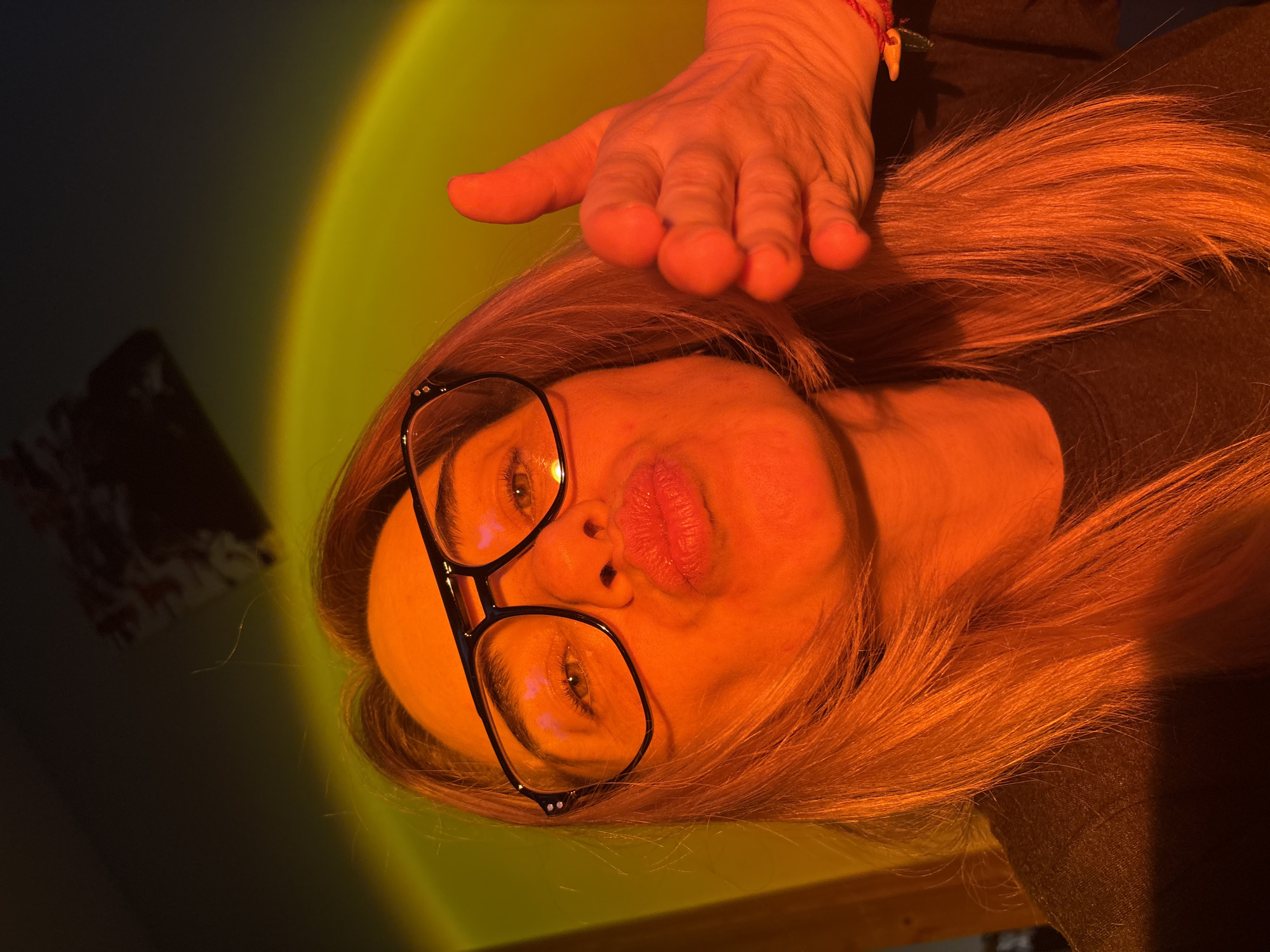An image captures a close-up of a person, likely female, posing indoors under warm, artificial lighting. The subject's head is tilted, and her face, framed by glasses, is central to the composition. She has long, reddish-blonde hair, which is illuminated by a strong orange and yellow glow originating from the upper left of the frame, possibly from a "sunset lamp" or similar light source.
The person's eyes are open, looking slightly upwards, and her lips are pursed in a "duck face" or blowing kiss expression. She wears dark-rimmed glasses. Resting on her head, partially obscuring her hair on the right side, is a human hand with fingers spread slightly. A red and beige bracelet is visible on the wrist of this hand.
The background on the left side is dark and indistinct, with faint, shadowy shapes that suggest furniture or other indoor objects. The right side of the background is dominated by the bright, circular gradient of the light source, transitioning from deep orange to yellow. No visible text is present in the image. The location is unknown, Russia.