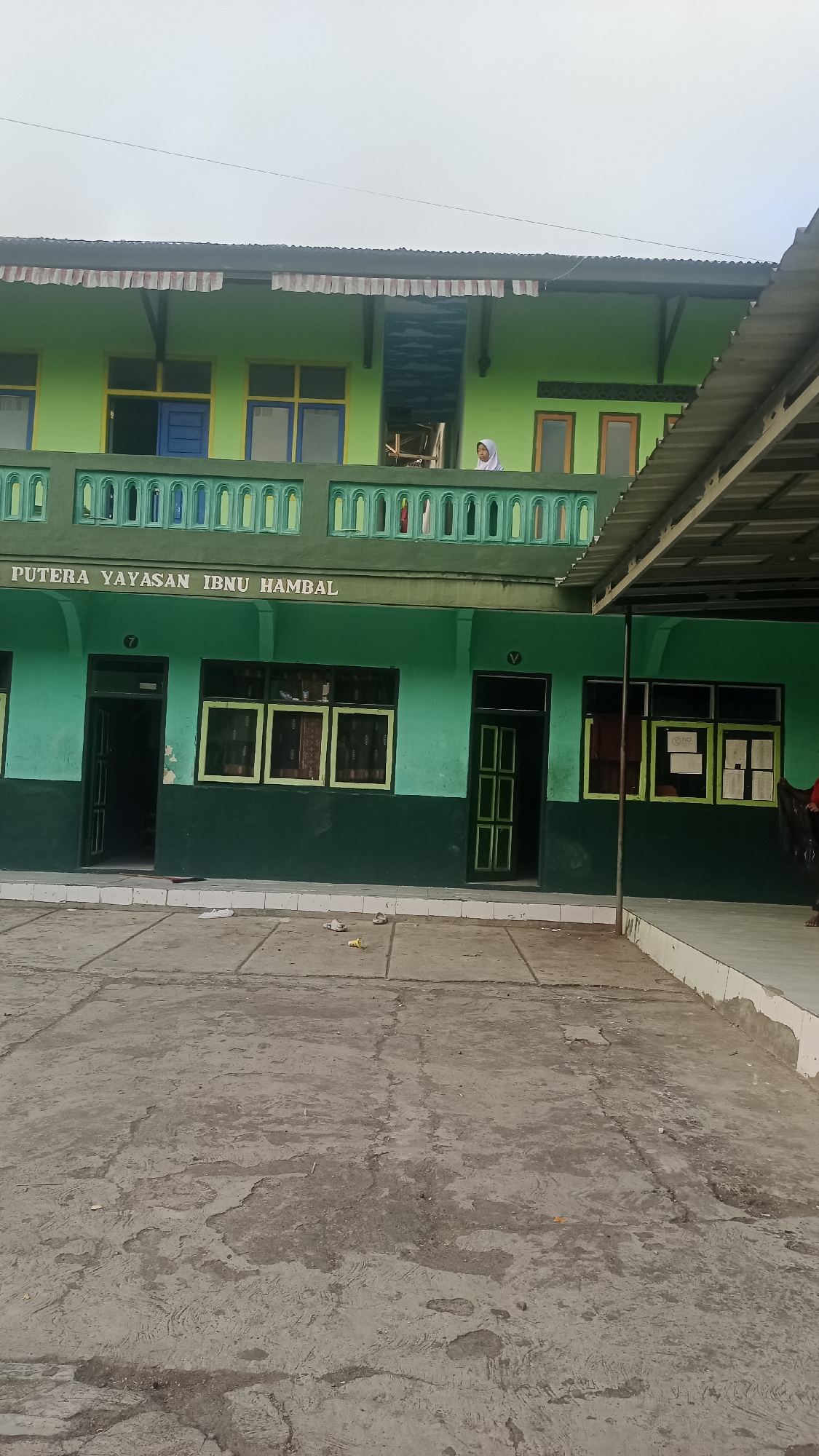The image shows the exterior of a two-story building, likely a school, with bright green walls. The building is located in the city of Karyamandala, Indonesia. On the first floor, there are several windows with yellow frames and a dark green base. Some doors are open, revealing the interior of rooms. The second floor also has windows with yellow and blue frames. A balcony with a green decorative railing runs along the second floor. A person wearing a white hijab is visible standing on the second-floor balcony. The foreground is a cracked concrete courtyard with some scattered debris, including what appear to be sandals. The sky is overcast. The text "PUTERA YAYASAN IBNU HAMBAL" is visible on the facade of the building.