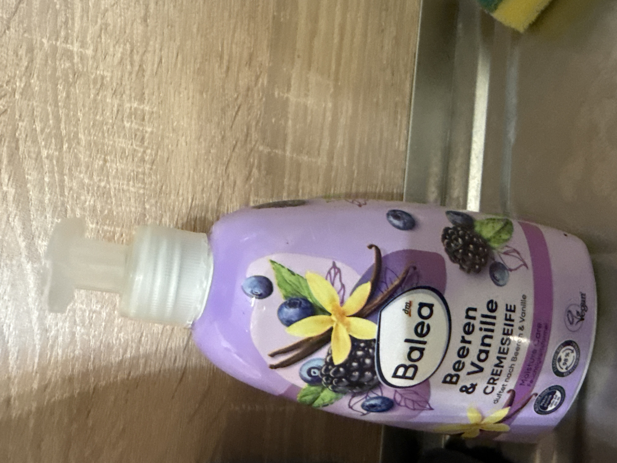A close-up, eye-level, medium shot shows a purple bottle of Balea "Beeren & Vanille CREmeseife" (Berry & Vanilla Cream Soap) with a white pump dispenser. The bottle is placed on a metallic surface, possibly a sink or countertop, with a wooden-paneled wall in the background. The bottle label features illustrations of blackberries, blueberries, and vanilla beans. Circular icons, including one indicating "98%," are visible at the bottom of the bottle. The overall lighting is dim. The image does not provide any visual cues to determine its location in Burgas, Bulgaria.