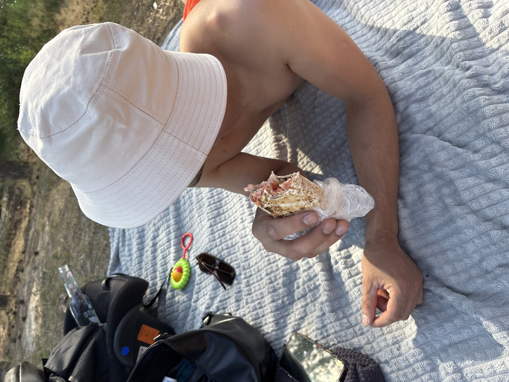 A person wearing a white bucket hat lies on a textured grey blanket outdoors, holding a wrapped food item in their hand. The person is shirtless and appears to be resting. Scattered around the blanket are several objects: a black backpack, a dark bag, sunglasses, a baby rattle, a glass bottle with the partial text "OTIM" visible, and a smartphone. The background suggests an outdoor environment with dry grass and vegetation, indicative of a daytime setting. The sunlight casts shadows on the blanket, suggesting it is either morning or late afternoon. The scene appears to be relaxed and casual, possibly a picnic or a break during an outdoor activity. The location context is Kurovskoye, Russia.