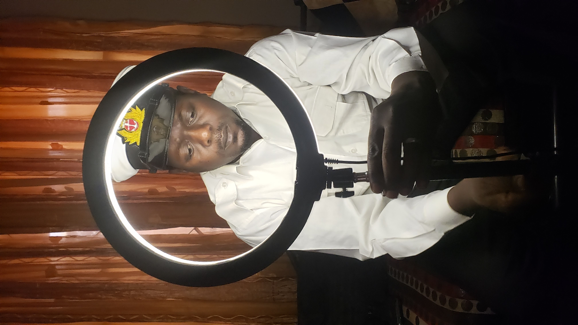 This media file captures a man indoors, illuminated by a large, circular LED ring light that frames his head and upper torso. The man has a dark complexion and a short beard, looking directly at the camera with a neutral expression. He wears a white, long-sleeved collared shirt with two buttoned breast pockets, and a dark peaked cap. The cap is adorned with a prominent emblem featuring a shield containing a red and white cross, encircled by elaborate gold-colored laurel-like branches.
His right hand is visible, holding onto the stand or mounting mechanism of the ring light. The background is dimly lit, revealing warm, earthy-toned striped or patterned fabric, possibly a curtain. To the right and below his arm, a decorative object, possibly a drum or pot, with horizontal red, white, and black stripes and circular accents, is partially visible. The scene suggests an indoor setting in Sabon Tasha, Nigeria, likely in the evening or at night, with the ring light serving as the primary light source for photography or video. The image itself is rotated 90 degrees clockwise. No discernible text is visible within the frame.