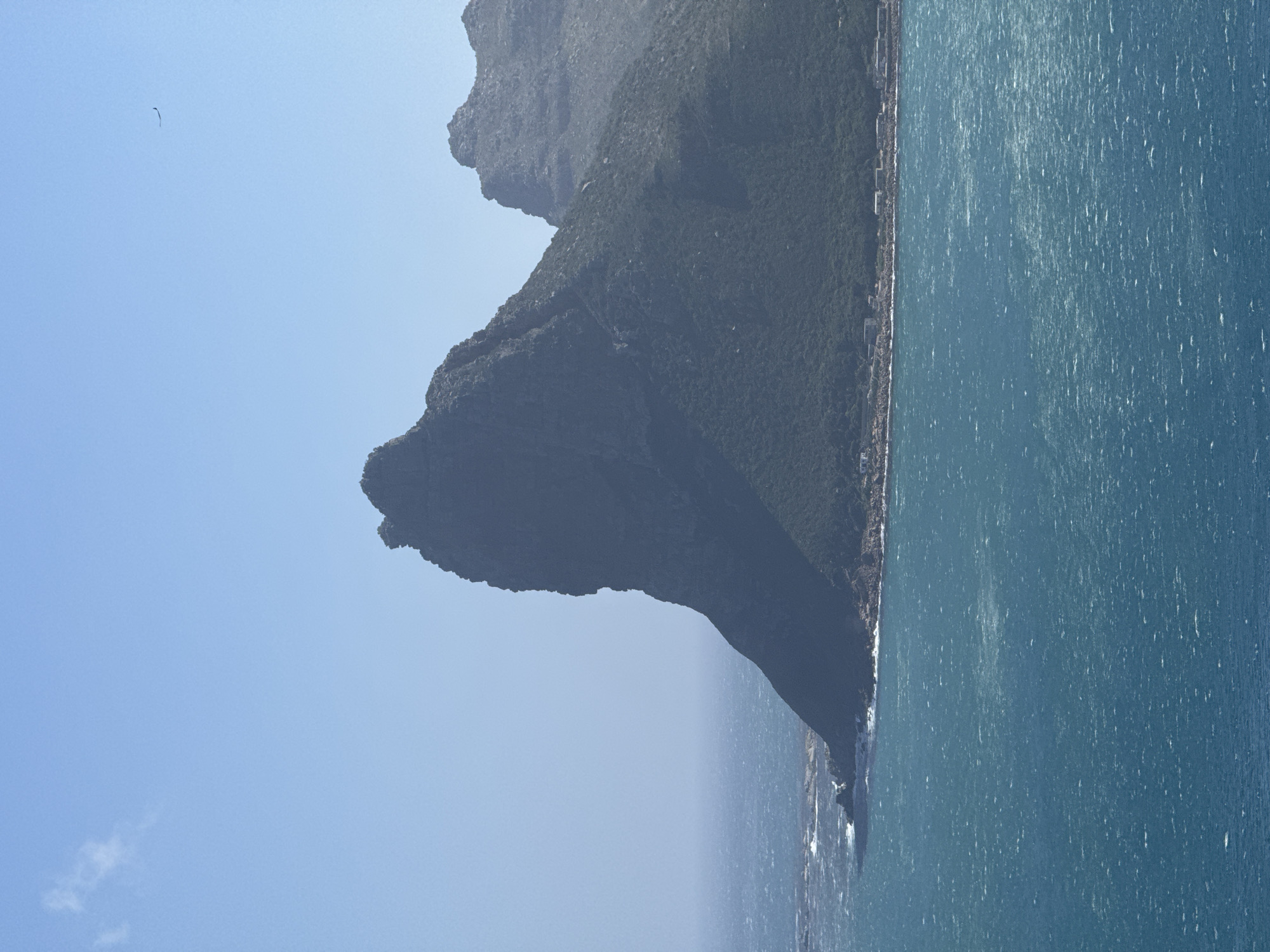 The image depicts a scenic view of the coastline near Hout Bay, South Africa, featuring a prominent, dark, rocky mountain formation jutting out into the turquoise ocean. The mountain is covered in dense, green vegetation, with a small cluster of buildings visible along its base, situated on a strip of land between the mountain and the sea. The ocean is choppy, with whitecaps indicating moderate wind. The sky is clear and blue, with a few wispy clouds and a distant, dark silhouette of what appears to be a bird or kite. The overall impression is one of a rugged, natural landscape meeting a coastal settlement under a bright, sunny sky.