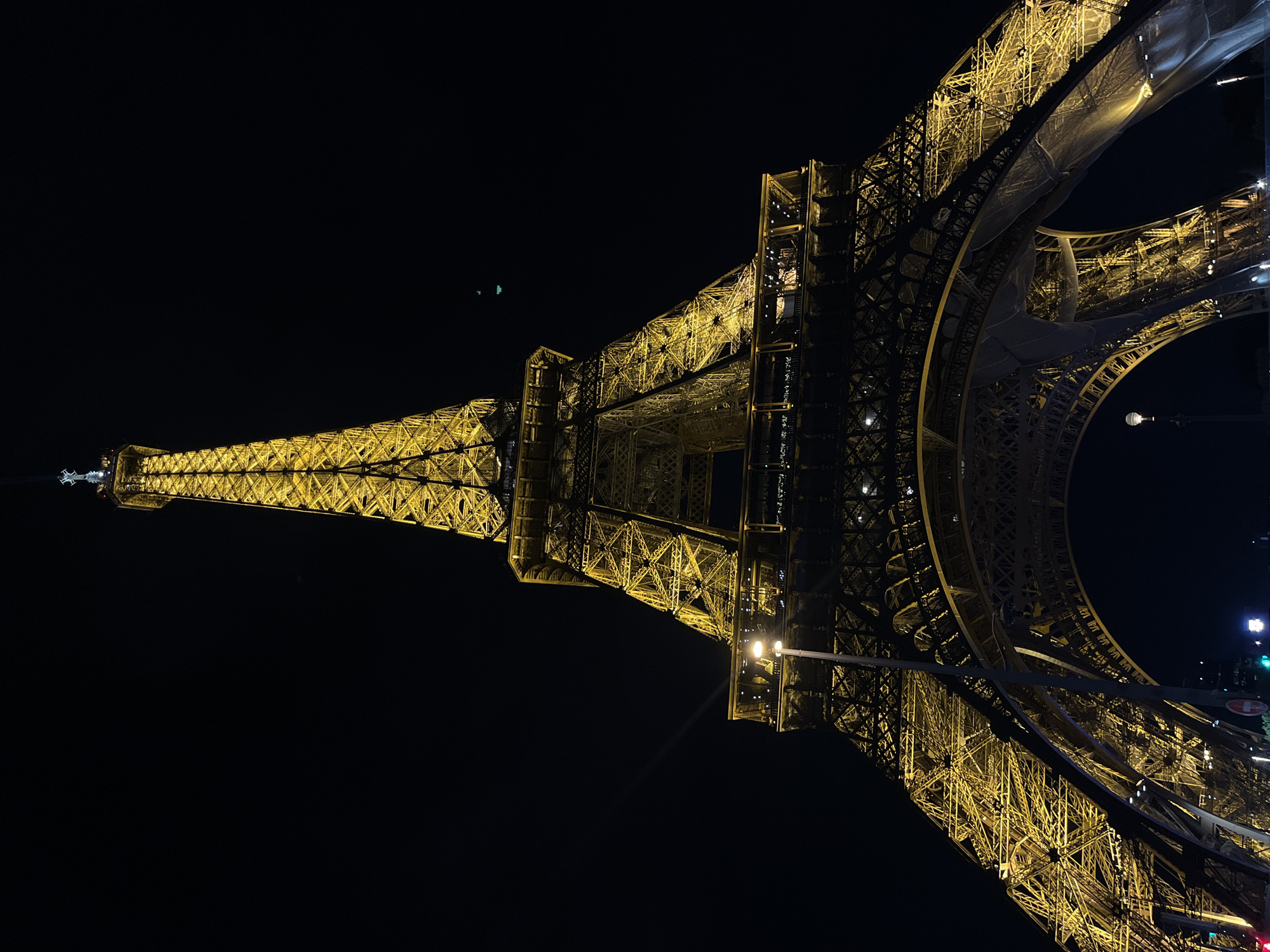 Illuminated against a black background, the Eiffel Tower in Paris, France is prominently displayed. The tower's intricate metal latticework is illuminated in a golden hue. A streetlight is present below the tower, and street lights are seen to the side. The shot is oriented vertically.