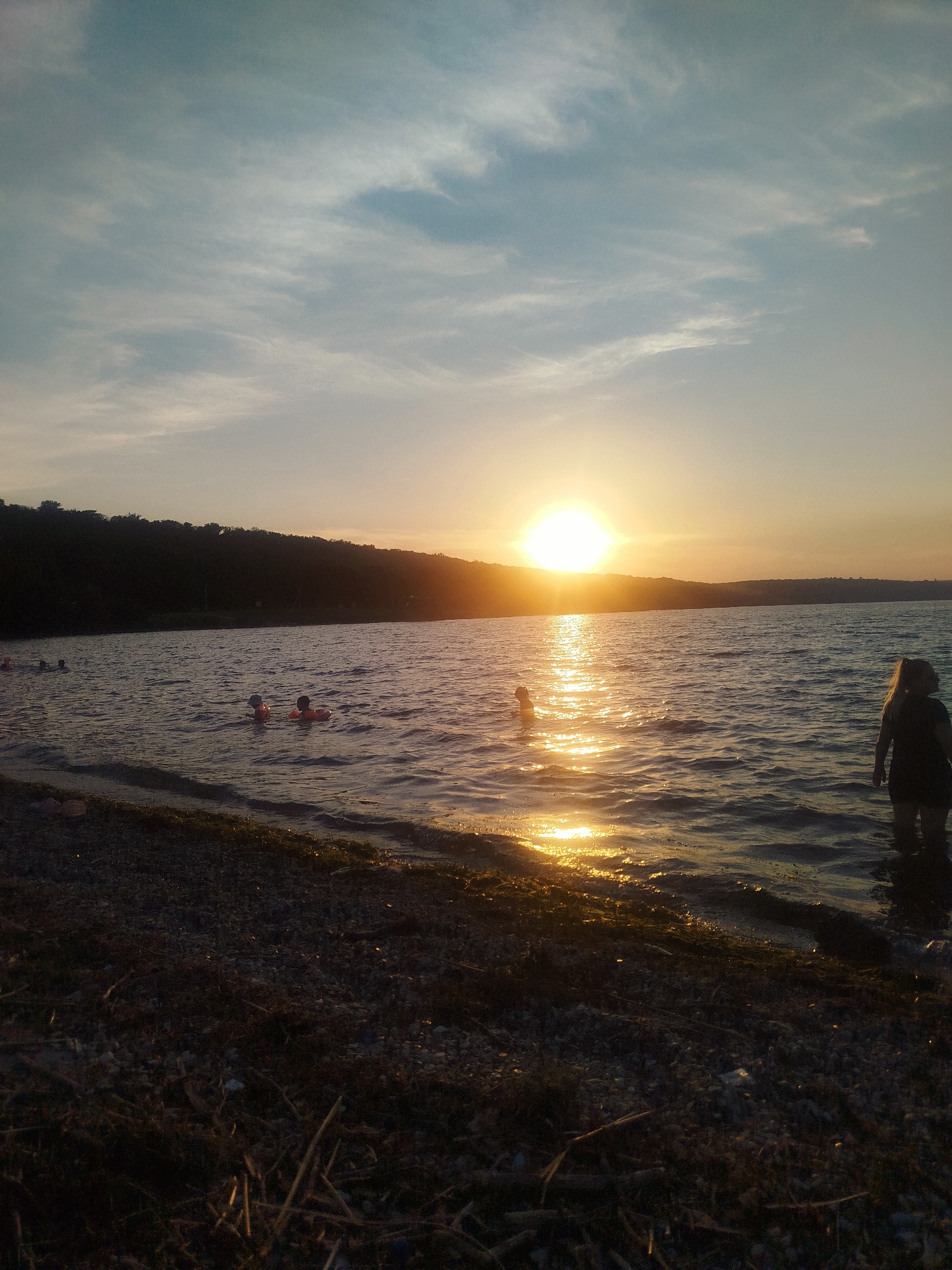 This media file captures a serene sunset scene at a body of water, likely a lake or river, near Ustia, Moldova. The sun is low on the horizon, casting a warm, golden glow across the water and the sky. Wispy clouds streak across the pale blue sky, tinged with the orange and yellow hues of the setting sun.
In the foreground, a pebbly shoreline is visible, with dried grass and twigs scattered on it. The water is gently rippling, reflecting the vibrant colors of the sunset. Several people are enjoying the water. In the distance, to the left, a few individuals are swimming. Closer to the center, two children are visible, each wearing an orange flotation device, suggesting they are young and learning to swim or playing in the water. Further to the right, a person stands at the edge of the water, facing away from the camera, their silhouette outlined against the bright sunset. The overall atmosphere is one of peaceful recreation and the beauty of nature at dusk. The time of day is clearly sunset, and the weather appears to be calm and pleasant. No visible text is present in the image.