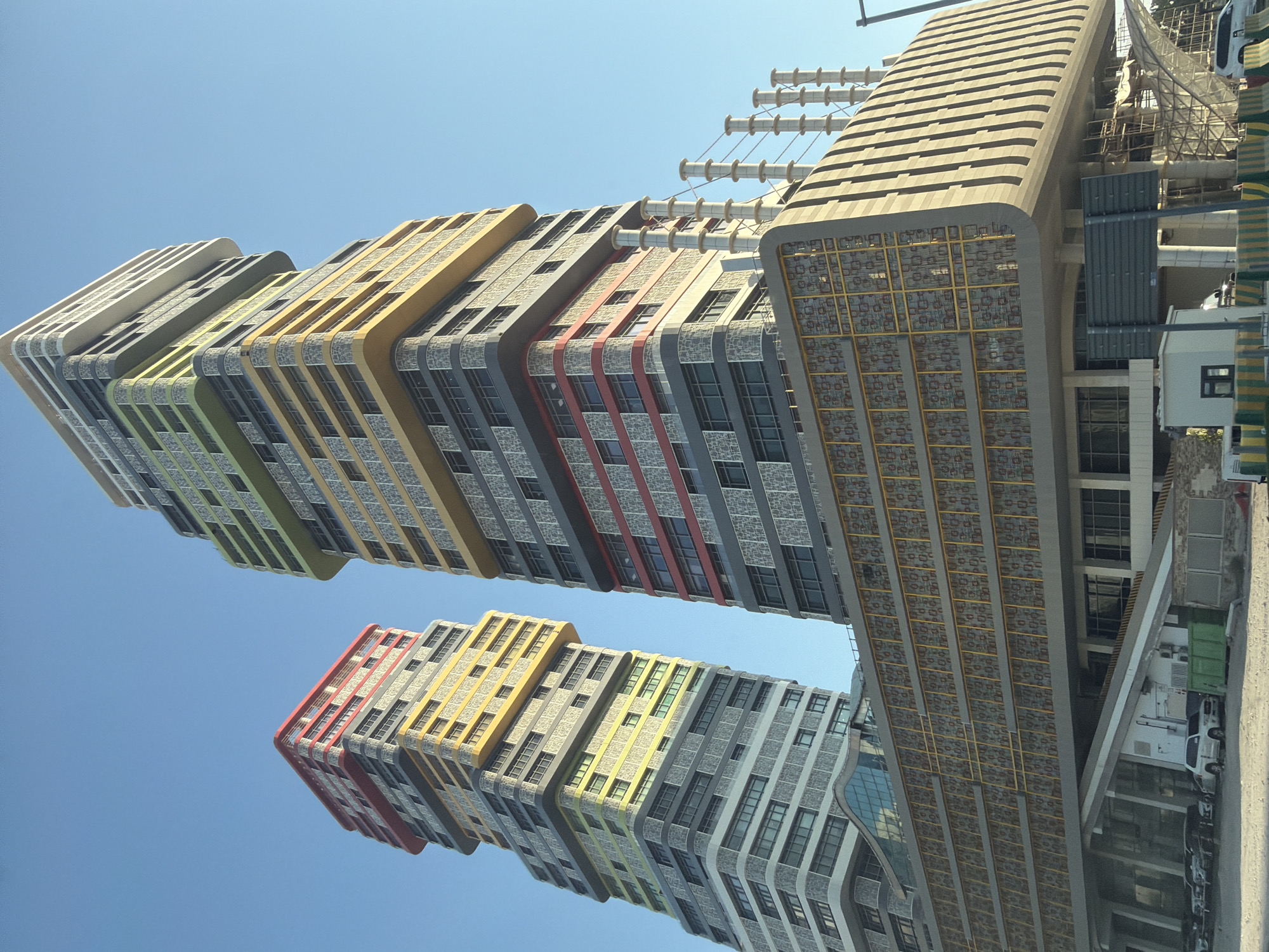 This media file shows a low-angle shot of several modern buildings against a clear blue sky. The buildings are tall and feature a distinctive, undulating design with colorful bands of yellow, green, red, and gray. They appear to be apartment buildings or offices, with numerous windows visible. One building on the right has a unique, textured facade with a geometric pattern and is partially under construction, with scaffolding and building materials visible. Another structure nearby appears to be a commercial building with large glass windows. A white vehicle and a green dumpster are also visible near the base of one of the buildings. The lighting suggests it is daytime, likely mid-morning or afternoon, with bright sunlight. The scene appears to be an urban environment, possibly a city under development. There is no visible text in the image.