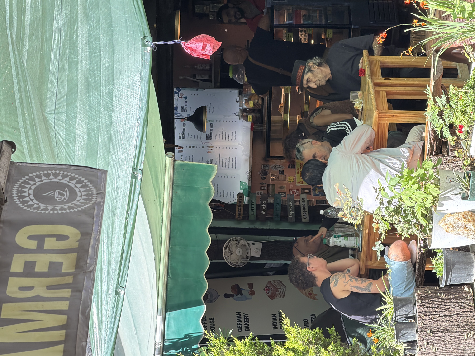 The image depicts a lively outdoor seating area at a cafe in Vashisht, India. A green awning provides shade over a wooden table where several individuals are seated. In the foreground, a person with tattoos on their arm, wearing a black sleeveless shirt and denim shorts, sits on a bench. To their right, two men converse, and further to the right, an elderly man with a distinctive hat and a grey beard is seated. Behind them, a busy cafe interior is visible, with a counter displaying food and drinks, and a menu board on the wall. The cafe appears to be a German bakery and cafe, with signage indicating "GERMAN BAKERY" and "COFFEE HOUSE." Various potted plants and trees surround the seating area, adding to the outdoor ambiance. The overall scene suggests a casual gathering and a moment of relaxation at a local establishment.
