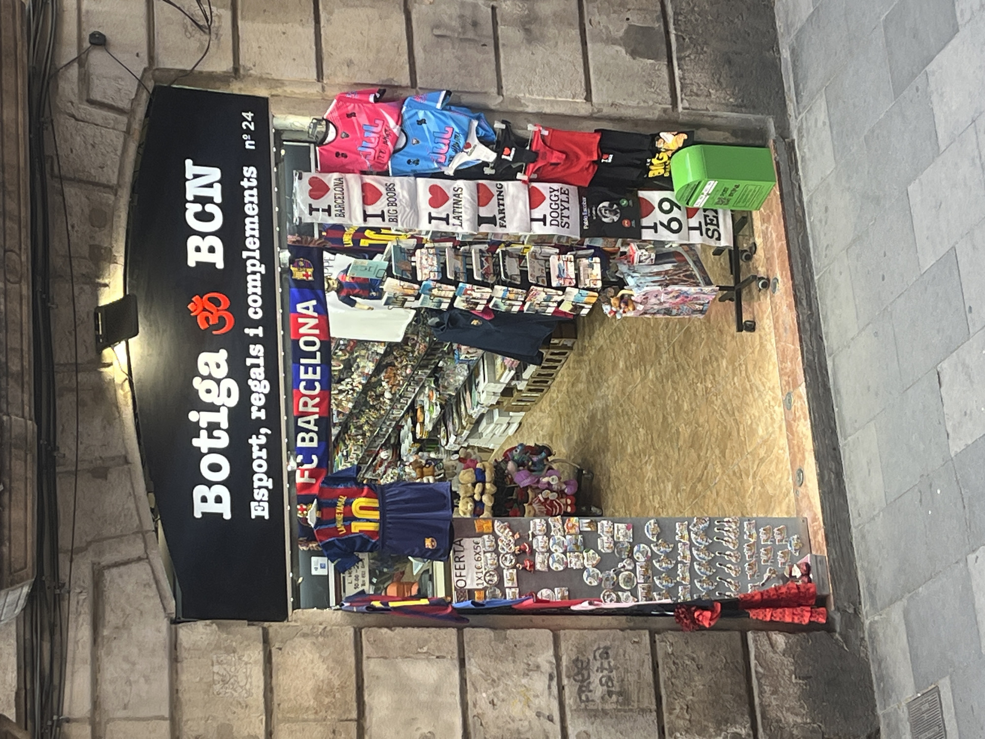 This image displays the vibrant entrance of a souvenir shop, situated in what appears to be a historic stone building in Barcelona, Spain. The shop's sign, oriented vertically, prominently features "Botiga ॐ BCN" in large white letters, with "Esport, regals i complements nº 24" (Sports, gifts, and accessories no. 24) below.
The storefront is packed with a diverse range of merchandise. Numerous t-shirts are displayed, including FC Barcelona jerseys, one clearly showing "LANGINE YAMAL 10". Adjacent to these are various novelty t-shirts with "I ♥" slogans, such as "I ♥ BARCELONA," "I ♥ BIG BOOBS," "I ♥ LATINAS," "I ♥ FARTING," "I ♥ DOGGY STYLE," and "I ♥ 69." Other t-shirts bear the text "JUL" and one reads "Pablo Escober." A green bin or box is visible near the t-shirt display.
Inside the shop, shelves are densely packed with more souvenirs, including stuffed animals and other trinkets. A display board filled with numerous pins or badges is positioned near the shop's entrance. A visible sign advertises an "OFERTA" (offer) stating "1X10.6X5€" and shop hours "10.00-22". The ground outside the shop entrance has "FREE" written as graffiti. The shop's offerings cater to both sports enthusiasts and those looking for humorous or quirky souvenirs. No people are visible in the shot, which is taken from a slightly elevated angle and rotated 90 degrees counter-clockwise.