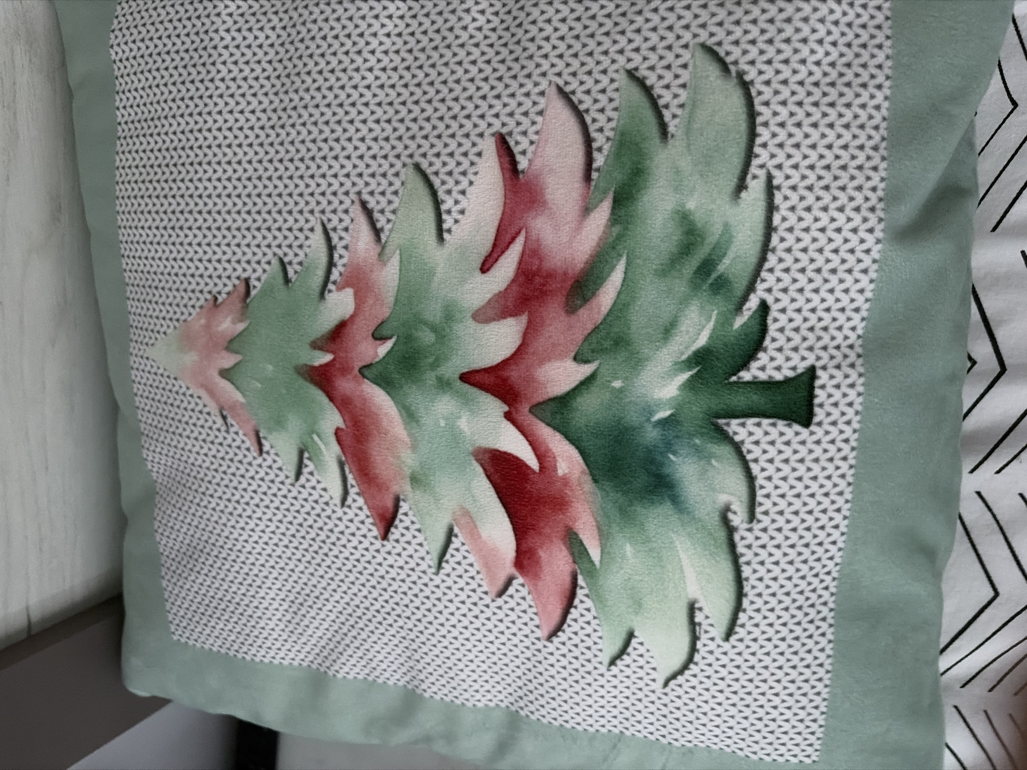 A close-up, overhead shot shows a decorative pillow with a Christmas tree design. The pillow is light green and has a textured gray and white knitted pattern. The Christmas tree is stylized, with red and green watercolor-like brushstrokes forming its branches. The tree has a small brown trunk and sits centrally on the pillow. The pillow is placed on a surface that appears to be a bed, with a white and black striped sheet visible at the bottom. The image does not contain any information about the city of Podgumer, Bulgaria.