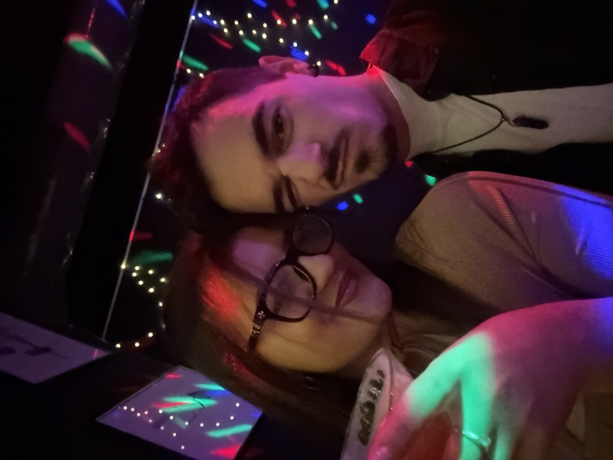 Two individuals, a female and a male, are captured in a close-up, informal portrait within a dimly lit entertainment venue, likely a bar or club in Sofia, Bulgaria. The specific location within Sofia is not discernible from the visual evidence.
The female, positioned on the left, has dark hair and wears dark-framed eyeglasses. She is dressed in a light brown top and holds a stemmed cocktail glass, possibly a martini, with dark garnishes. Her expression is a subtle smile directed towards the camera. Her right hand, holding the drink, features a ring and is illuminated by green and magenta light.
The male, on the right, has dark hair, a mustache, and a short goatee. He wears a white t-shirt beneath an open dark button-up shirt and a dark pendant on a cord necklace. A dark stud earring is visible in his left ear. He is making a playful expression, winking with his right eye and exhibiting a smirk.
The background is dark, adorned with numerous small, multi-colored lights in patterns, predominantly red, green, blue, and white/yellow, creating a festive atmosphere typical of a nightlife establishment. Reflections and patterns from these lights are visible on surfaces behind the subjects and cast colored hues onto their faces and clothing.