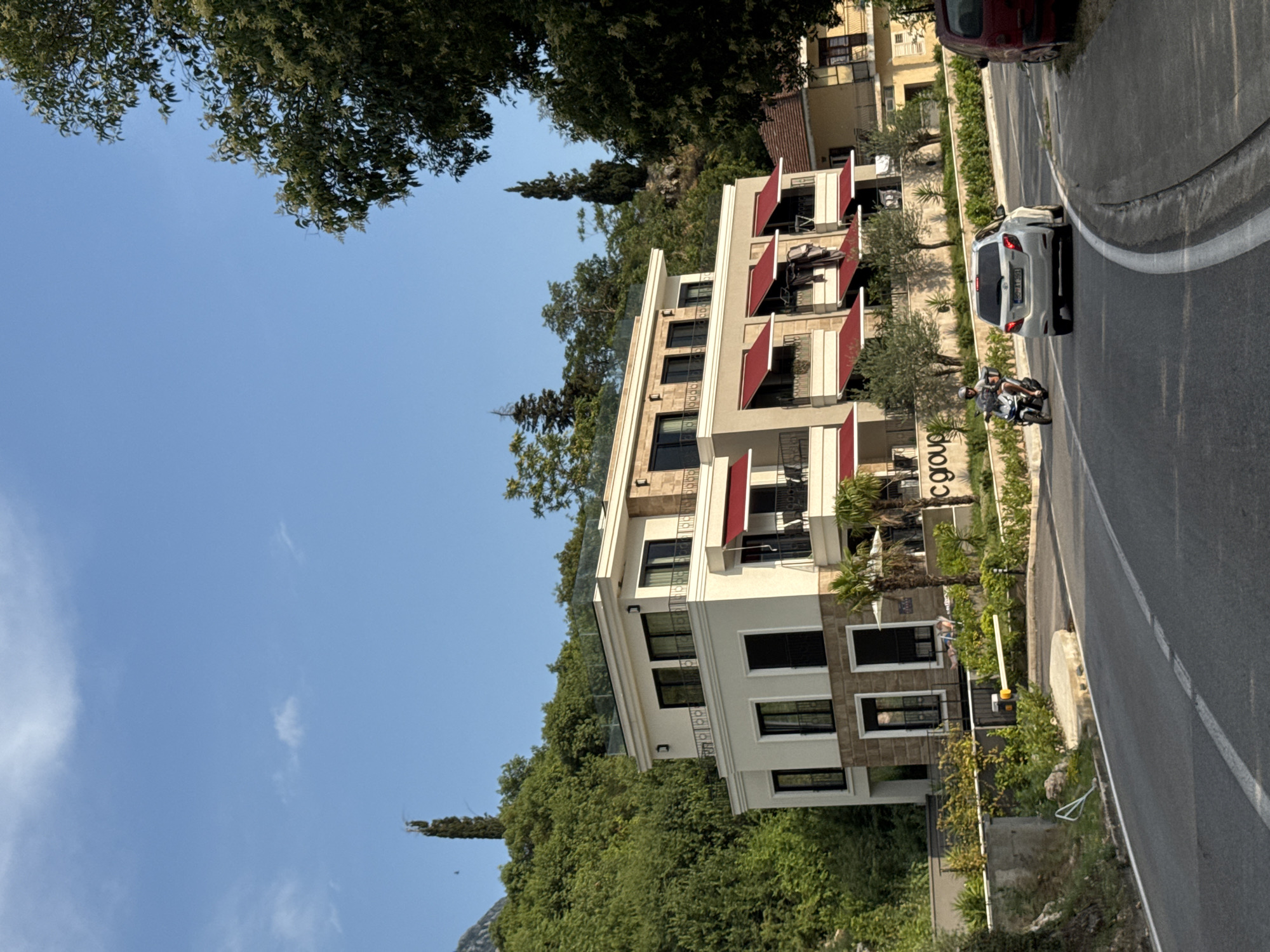 The image shows a modern white building with multiple levels, adorned with red awnings over balconies. The building is situated on a hillside covered in lush green trees, characteristic of Dobrota, Montenegro. In front of the building, a road curves to the right, with a white car driving on it. A person on a scooter is also visible on the road, moving away from the camera. The sky is clear blue with a few wispy clouds. The scene appears to be a sunny day in a scenic, coastal town.