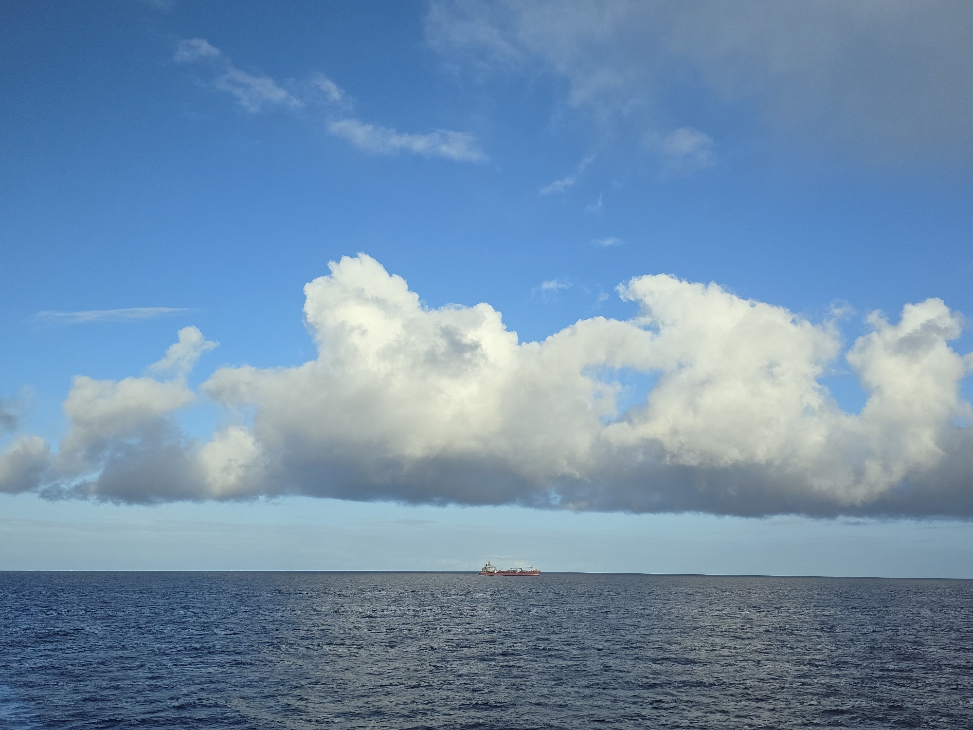 The image shows a body of water, presumably the ocean, under a bright blue sky with scattered white clouds. A cargo ship is visible in the distance, appearing small against the vast expanse of the water. The location is unknown.