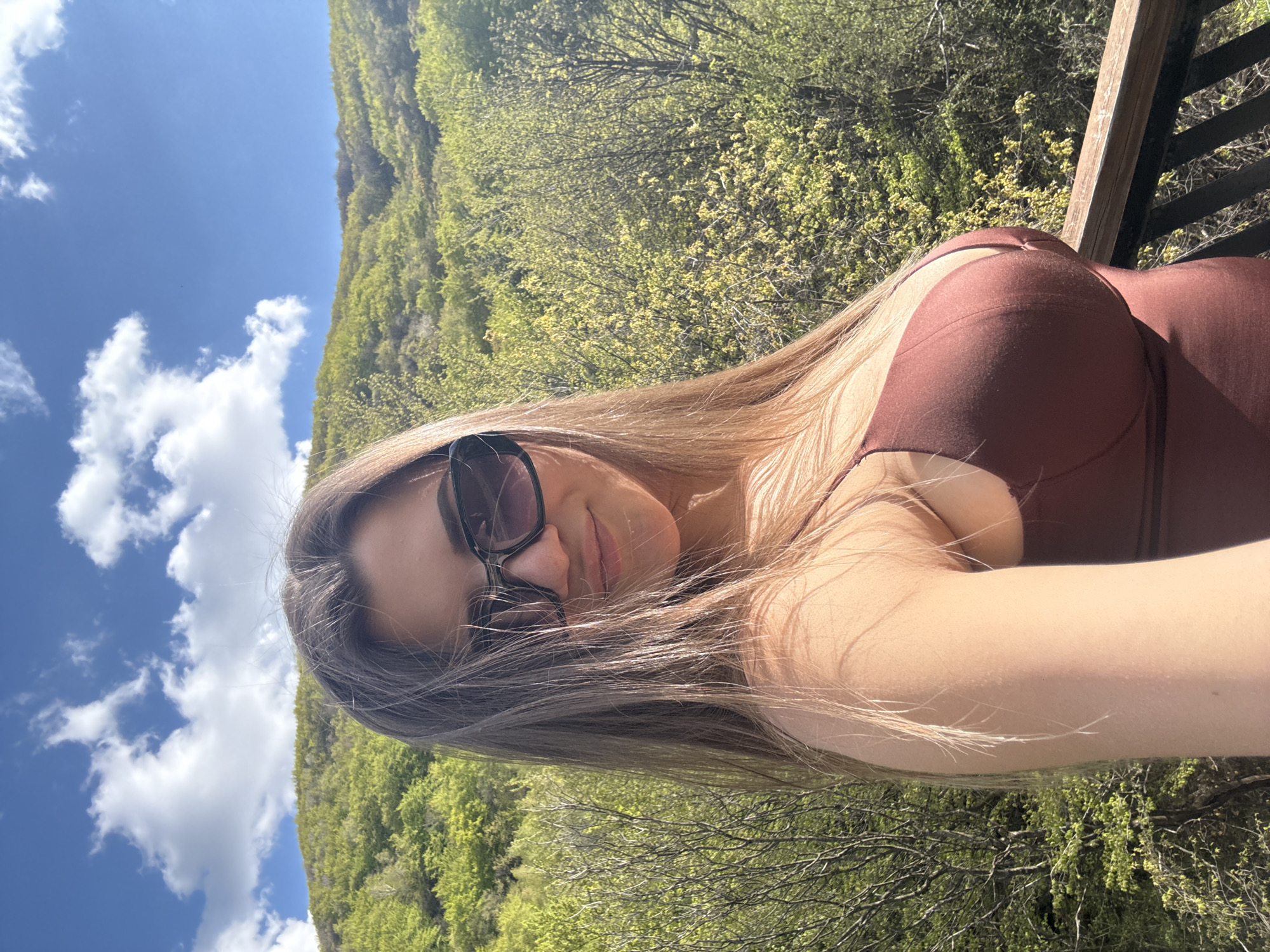 A woman with long brown hair is taking a selfie outdoors. She is wearing sunglasses and a brown top. Behind her, there is a forested hillside and a blue sky with white clouds. There is a wooden structure to her right. The exact location in Sofia, Bulgaria, cannot be determined from the given image.