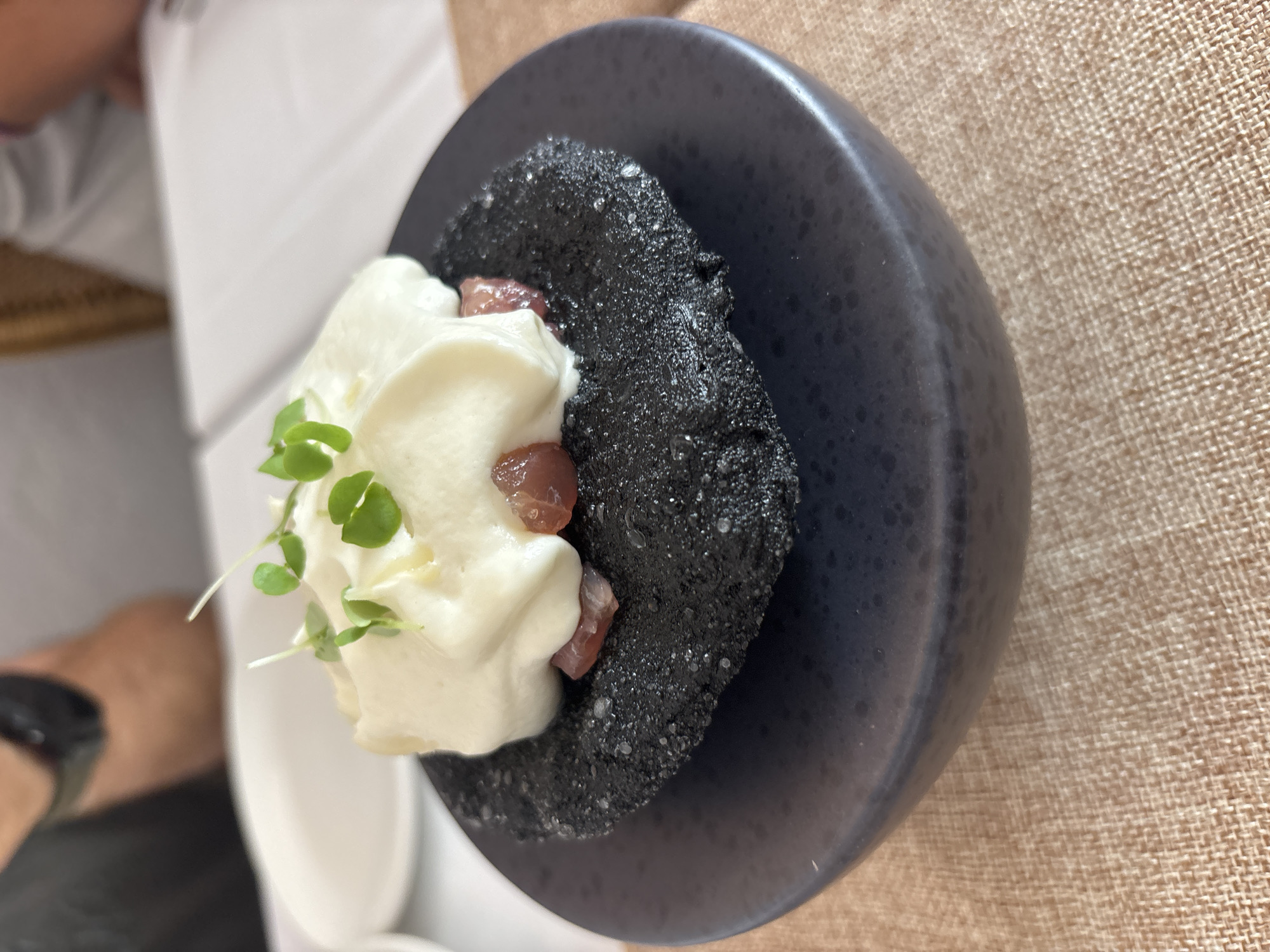 A close-up shot focuses on a small, dark, circular pastry topped with a dollop of white cream and garnished with microgreens. The pastry appears to be made from a dark flour or dough, possibly charcoal-infused, with a slightly rough texture. Beneath the cream, small pieces of red, finely diced food, likely tuna or another type of fish, are visible. The entire dish is presented on a dark, shallow plate, which sits on a textured, light brown placemat. In the blurred background, two individuals are seated at a table with white tablecloths, suggesting a dining setting. One person on the left is wearing a black t-shirt and appears to be looking down. The other person on the right is wearing a light-colored shirt. The setting is presumed to be in Carboneras, Spain, a coastal town known for its seafood restaurants, which would be consistent with the presentation of a dish featuring raw fish.