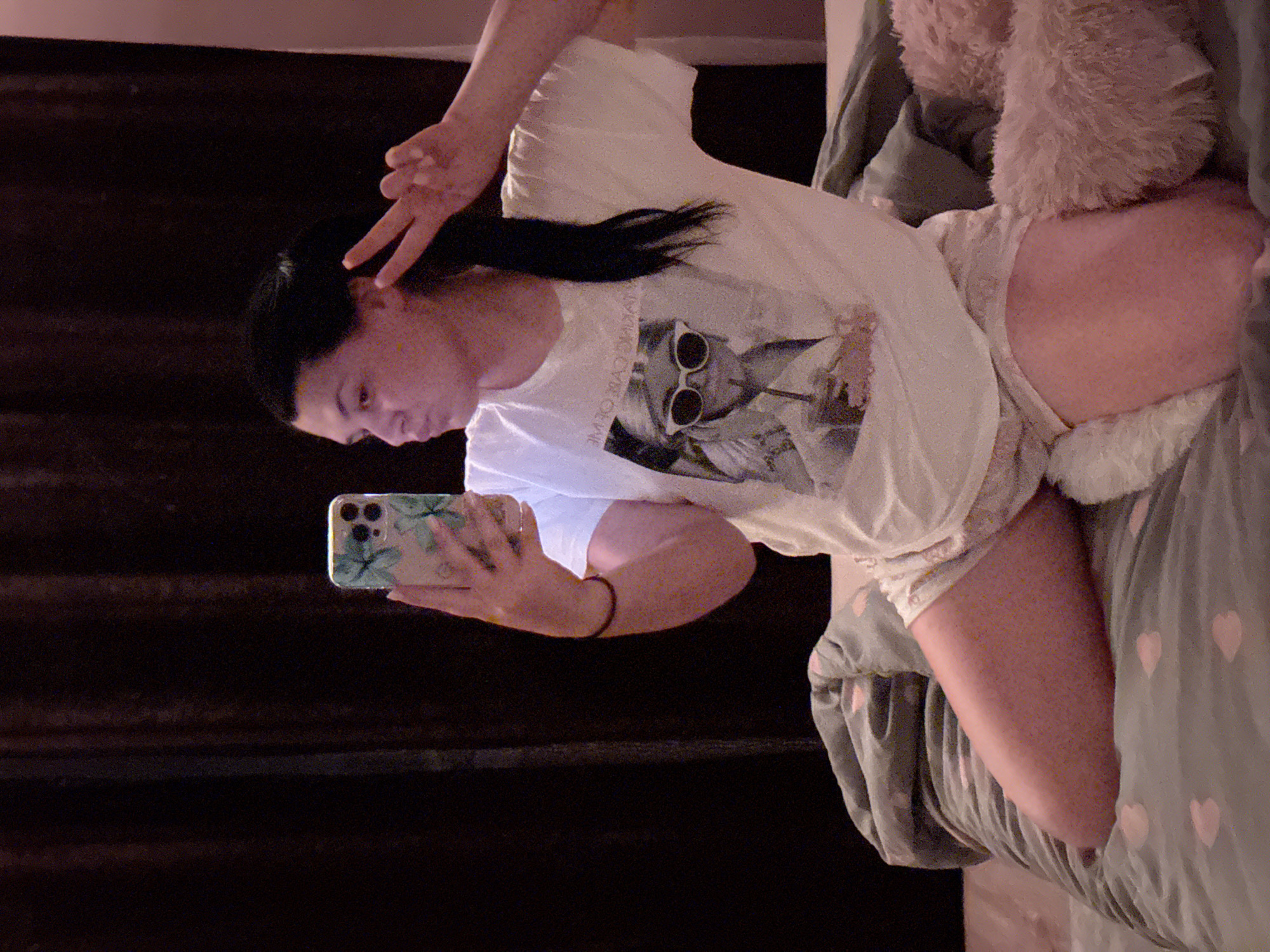 A person is depicted lying on a bed, looking towards a smartphone held in their left hand. The image is oriented vertically, showing the person lying sideways. They have dark hair, pulled back, and are wearing a white t-shirt with a graphic print of a cat wearing sunglasses. The graphic also contains white, mirrored text that appears to read "WILD CAT CREW." The person is also wearing light-colored shorts. Their right hand is raised, forming a peace sign or "V" gesture.
The setting appears to be a bedroom, with a grey-green duvet cover featuring pink heart patterns visible on the bed. A fluffy, light-colored pillow is seen near the top right of the frame. The background is dark, suggesting indoor nighttime or evening lighting. The smartphone case is light-colored with a green and blue floral or abstract pattern. The person's expression is neutral, and the overall mood is relaxed and possibly playful.