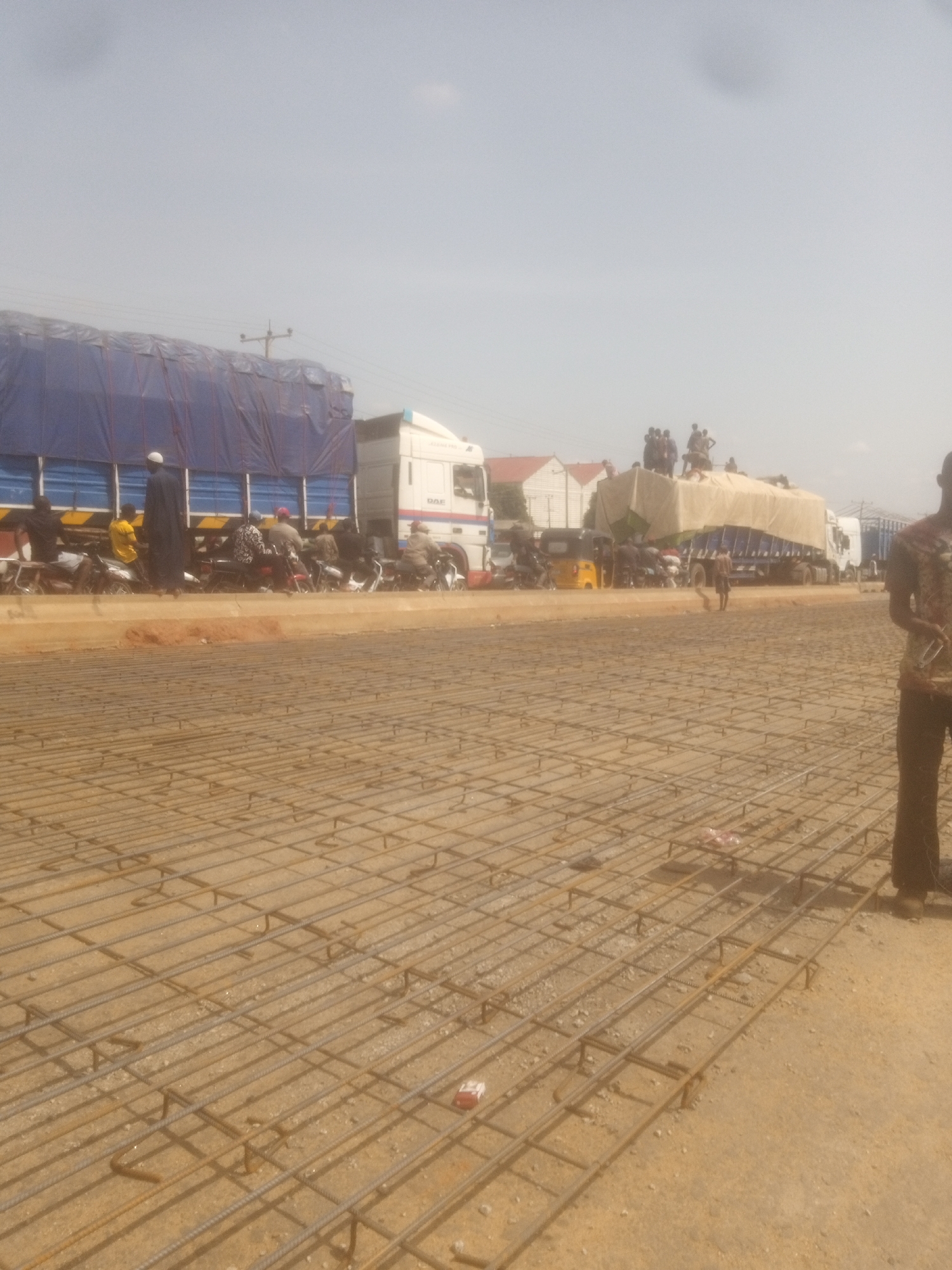 The image depicts an outdoor scene in Nassarawa, Nigeria, likely on a construction site or a road being prepared for paving. In the foreground, a grid of steel rebar lies on the ground, indicating the initial stages of concrete work. The rebar is laid out in a regular pattern, suggesting preparation for a large surface, possibly a road or a foundation.
In the middle ground, several large trucks are parked, some of them covered with blue tarpaulins. One truck appears to be a DAF model. There are groups of people, some standing on top of the trucks, and others are seen near the vehicles, with some on motorcycles and a yellow three-wheeled auto-rickshaw. The presence of these vehicles and individuals suggests activity related to transportation and logistics, possibly involving the delivery of materials or people.
In the background, there are some buildings, possibly houses, with red roofs. The sky is hazy or overcast, with a light cloud formation visible. The overall environment appears to be dusty and arid, consistent with a Nigerian setting. On the right side of the image, a person in dark clothing is standing, partially visible from the waist down, suggesting their involvement or observation of the ongoing activities. The scene is brightly lit, indicating daylight.