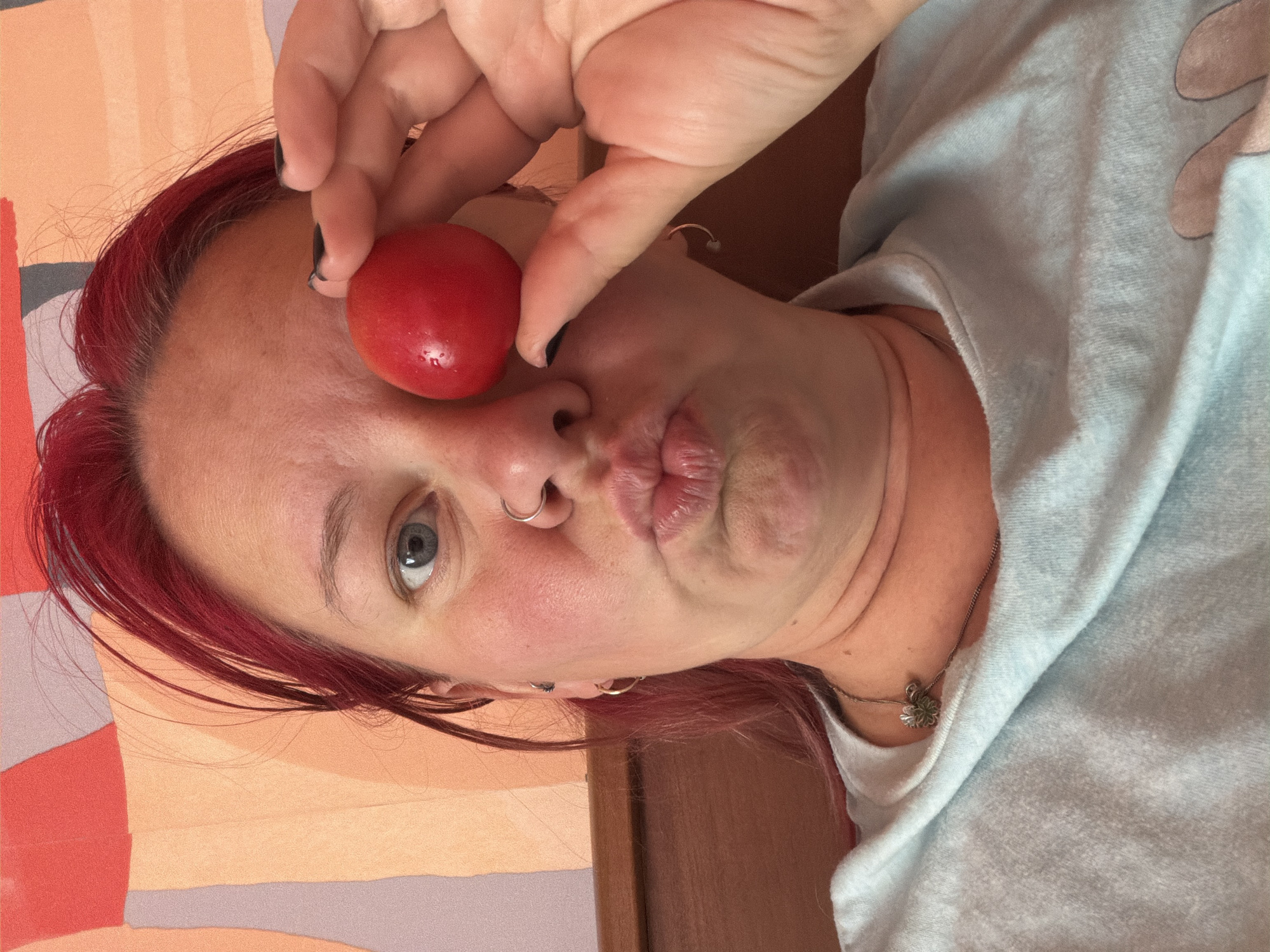 A close-up, selfie-style portrait features a woman with bright red hair and blue eyes. She is holding a single, round, red fruit, possibly a cherry tomato, in front of her right eye, obscuring it partially. Her lips are pursed, creating a comical expression. She is wearing a light blue t-shirt with a silver necklace featuring a clover-like pendant. The background consists of a wall with an abstract pattern of colored stripes in shades of peach, pink, and grey. The image appears to be taken indoors, possibly in a bedroom, given the presence of a wooden headboard and a soft, textured wall covering. The lighting is natural and casts a gentle glow on the subject's face. There is no indication of the city of Chaikovsky, Russia.